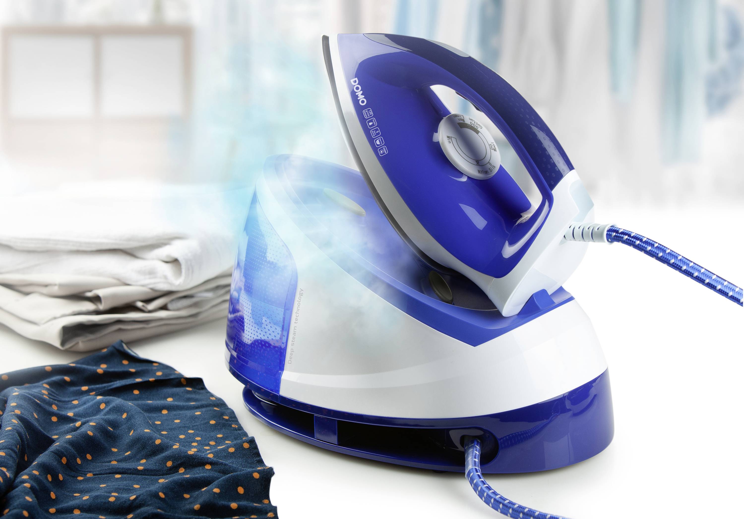 Un fer à repasser bleu et blanc sur une planche à repasser, entouré de vêtements repassés et non repassés. Des vêtements flous en arrière-plan.