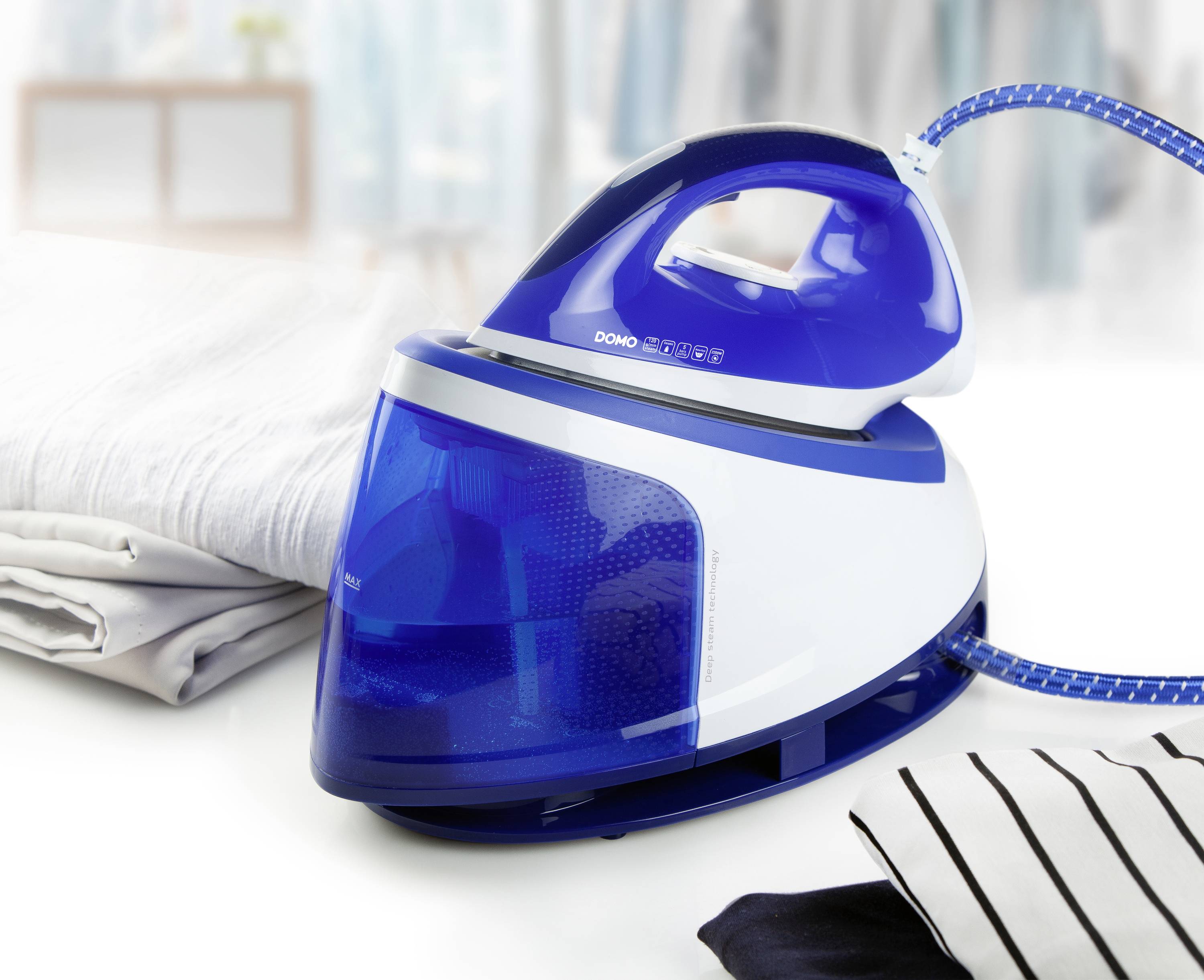 Un fer à repasser vapeur bleu et blanc est posé sur une planche à repasser à côté d'une pile de chemises et de chemisiers repassés, avec un arrière-plan flou.