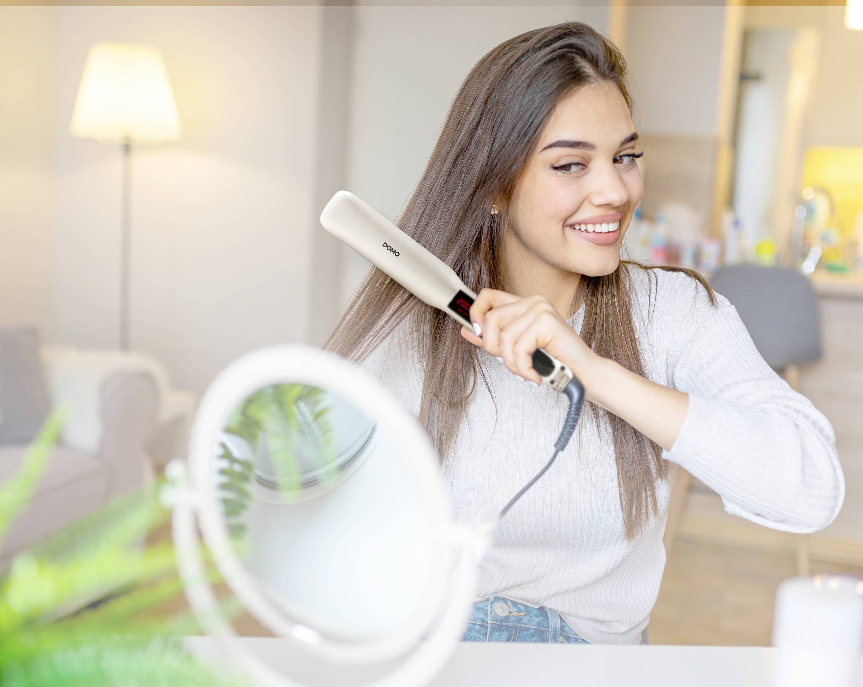 Une femme lisse ses longs cheveux bruns avec un fer à lisser dans une pièce confortable avec une lampe et un miroir. Elle sourit pendant qu'elle le fait.