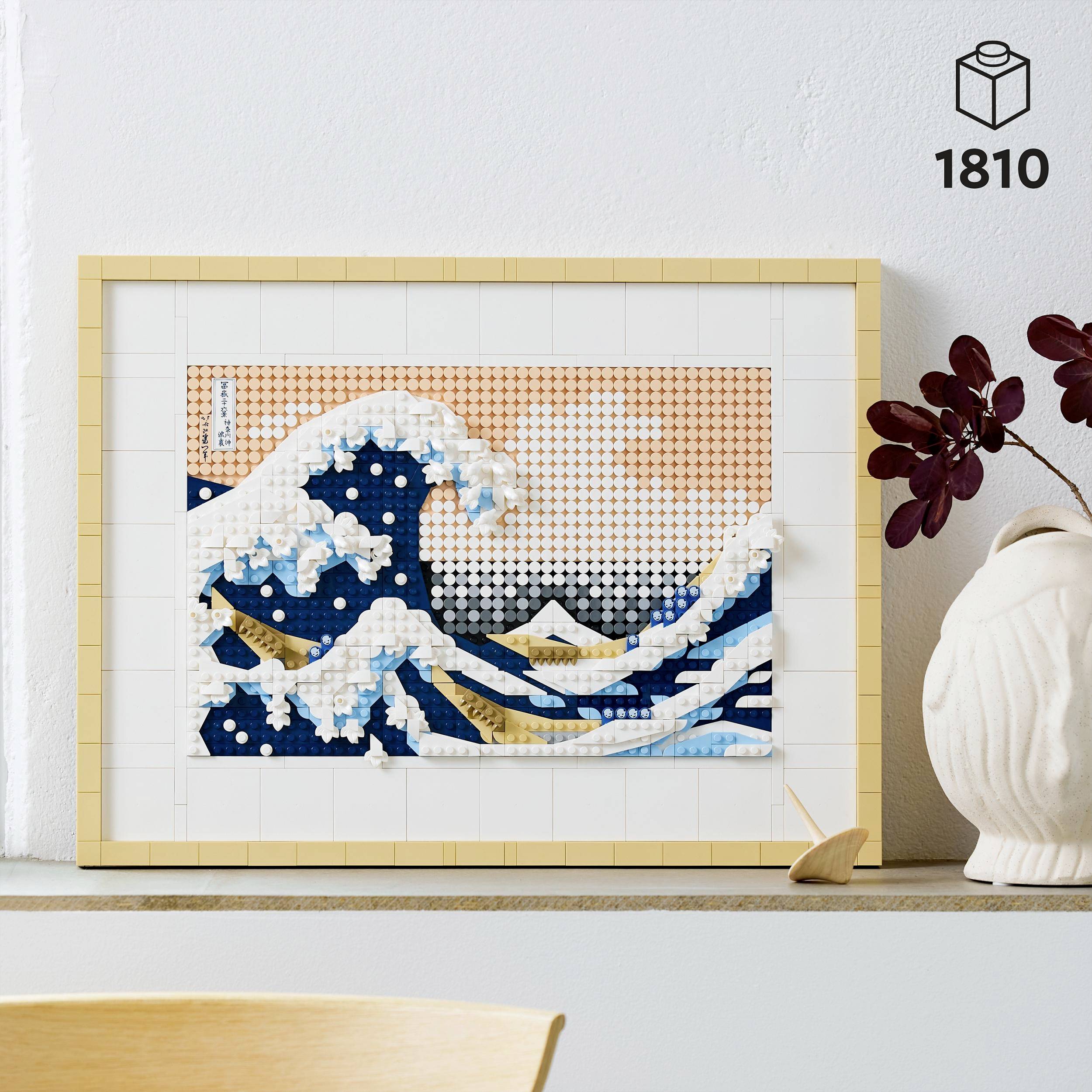 Une image LEGO encadrée représente un paysage ondulant inspiré de 'La Grande Vague' de Hokusai. Elle est posée sur une étagère à côté d'un vase.