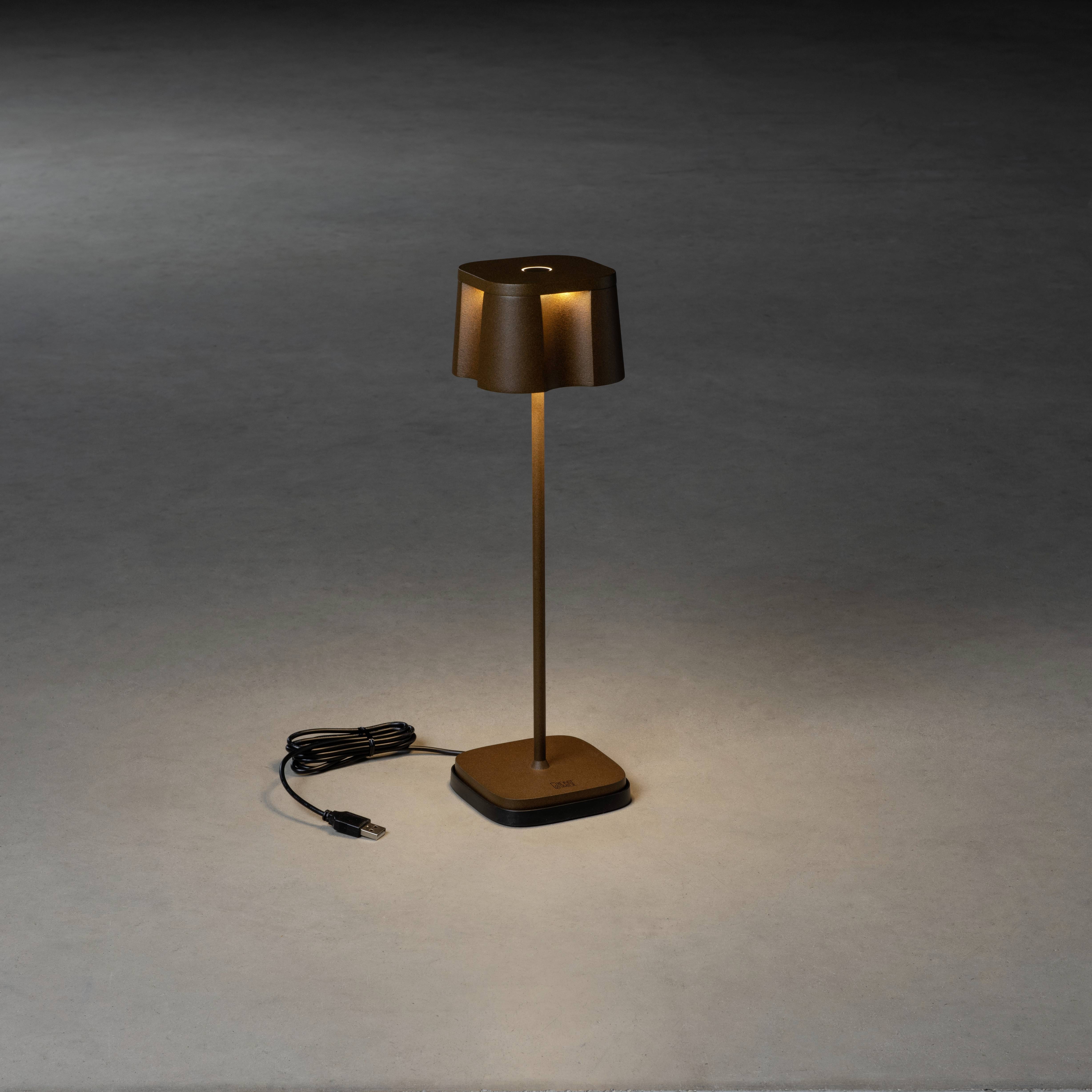 Une lampe fine et brune avec un abat-jour rectangulaire est posée sur un sol sombre, le câble électrique étant enroulé à côté.