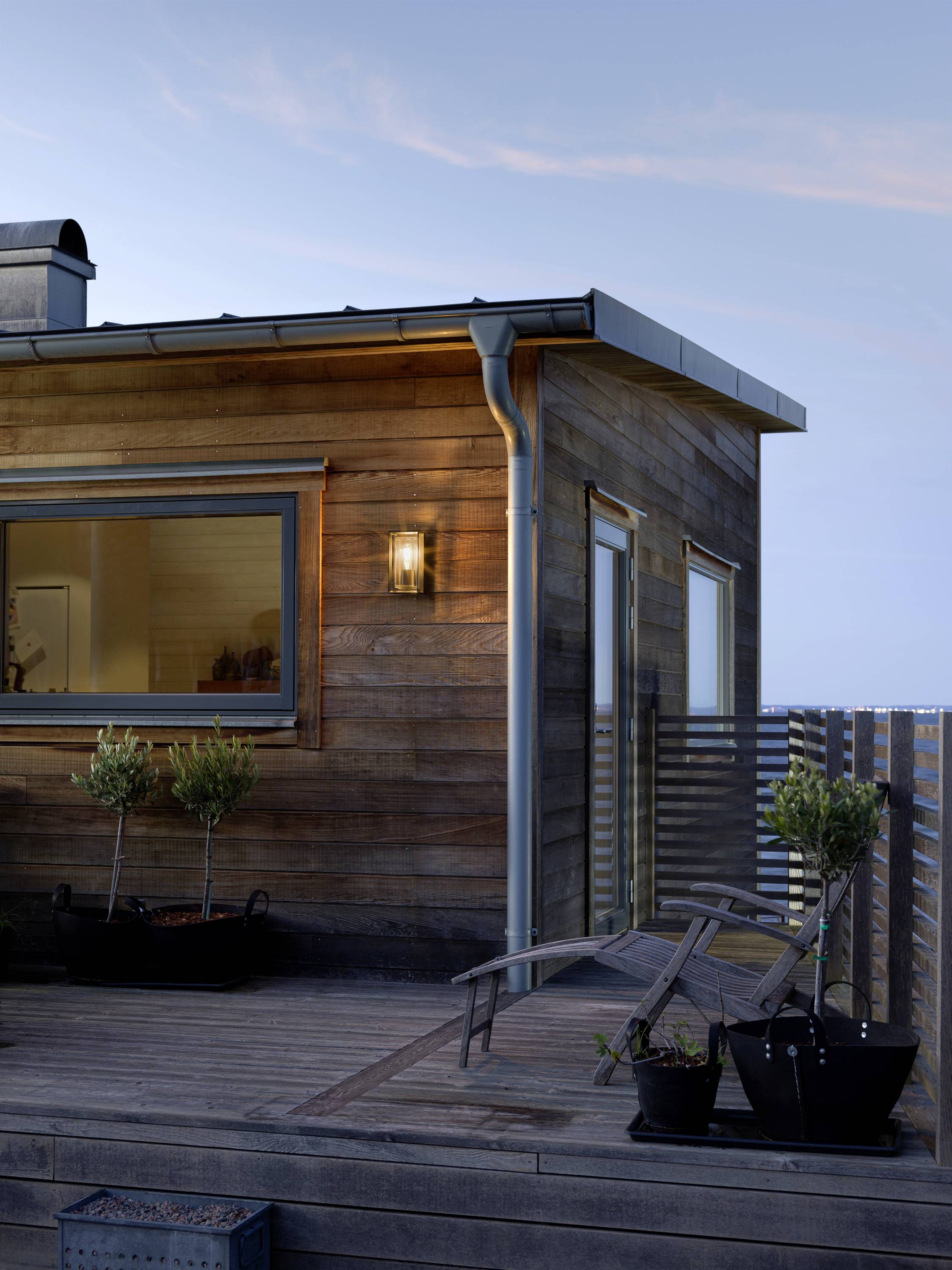 Une maison en bois moderne avec une fenêtre éclairée et une terrasse où se trouvent des plantes et un transat, dans un décor de soirée.