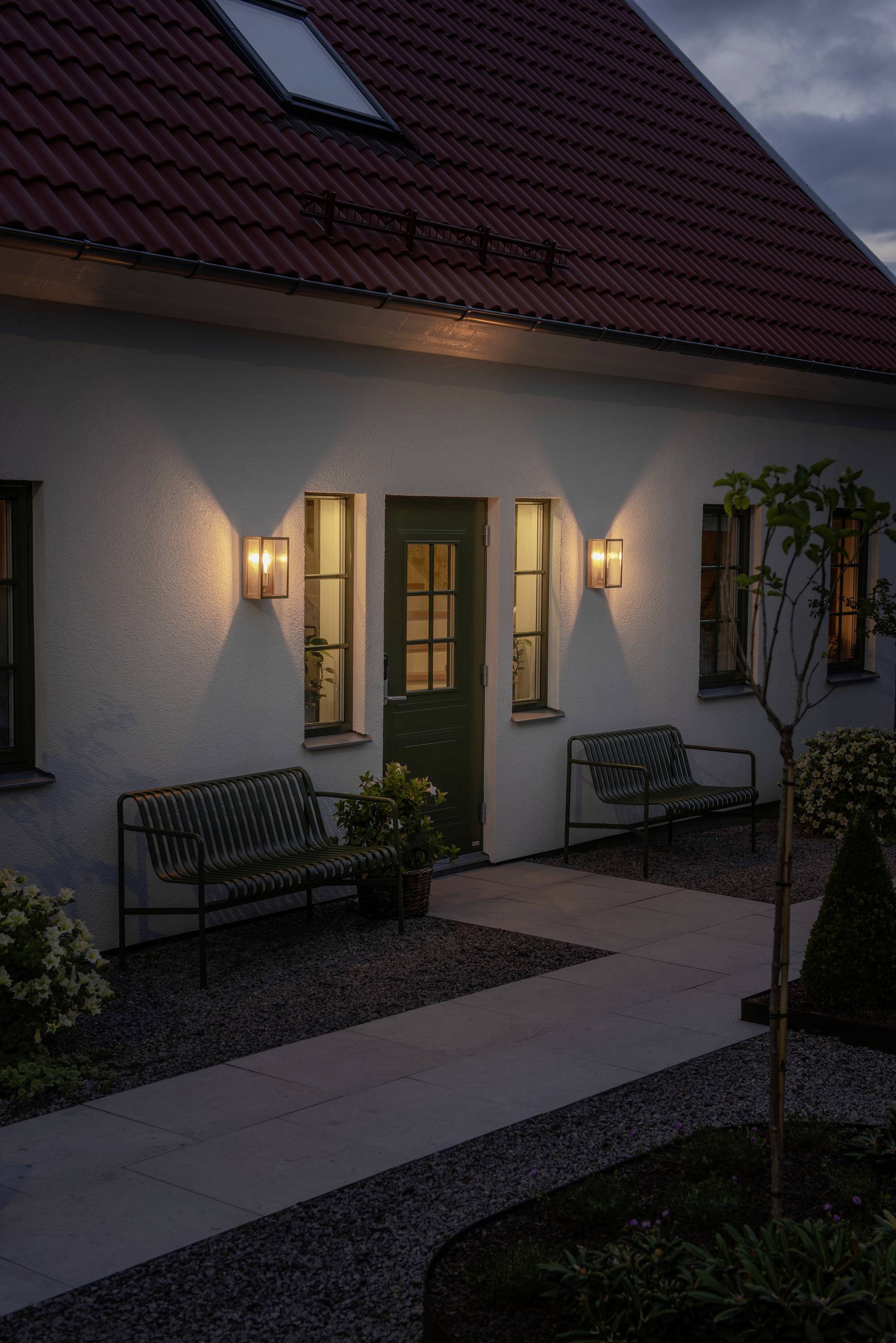 Une maison illuminée au toit rouge à la tombée du jour, sa porte d'entrée encadrée par deux lampadaires extérieurs allumés. Deux bancs de jardin sont disposés devant.