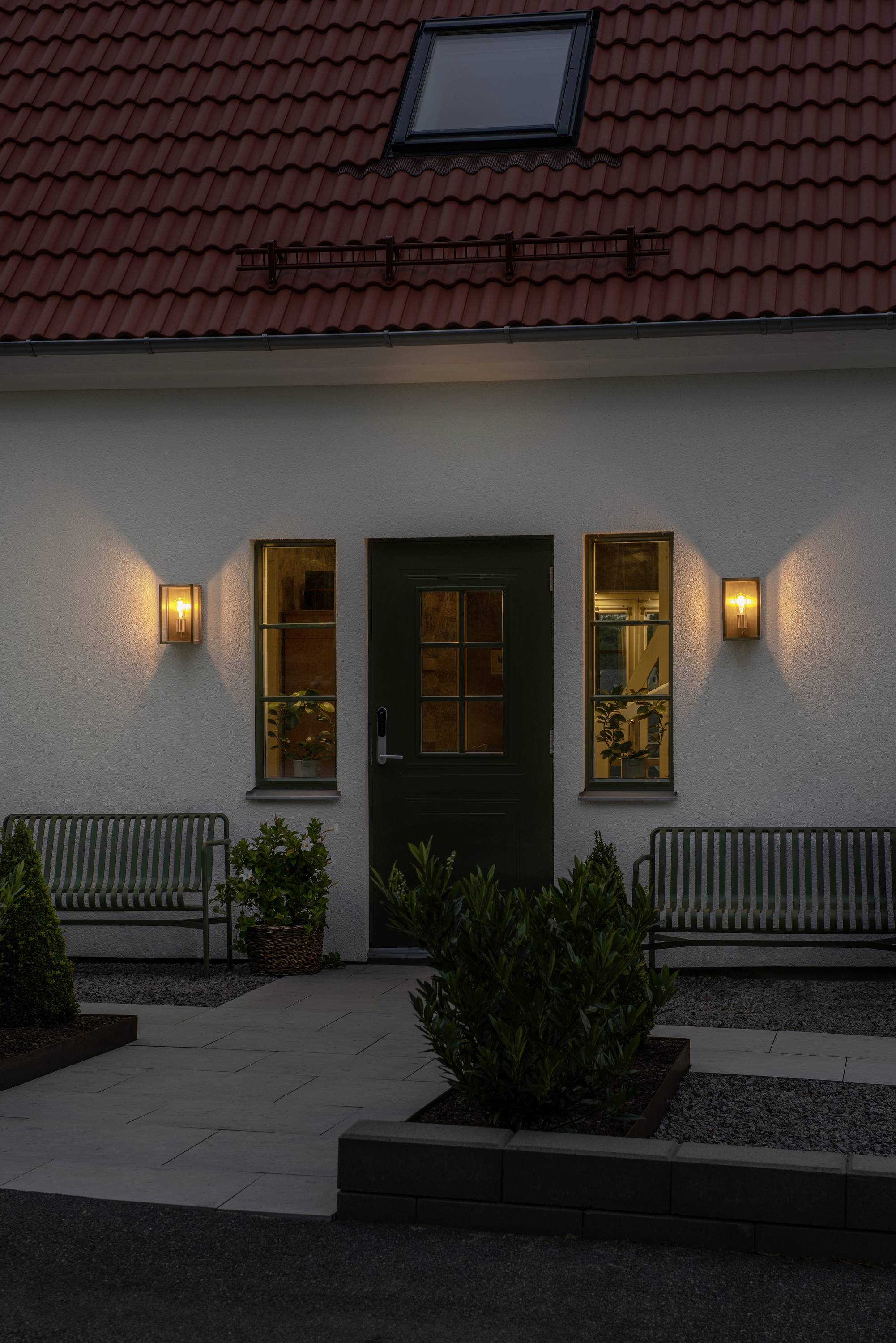 Entrée d'une maison avec une porte verte et des appliques murales éclairées. Deux bancs sont placés de part et d'autre de la porte.