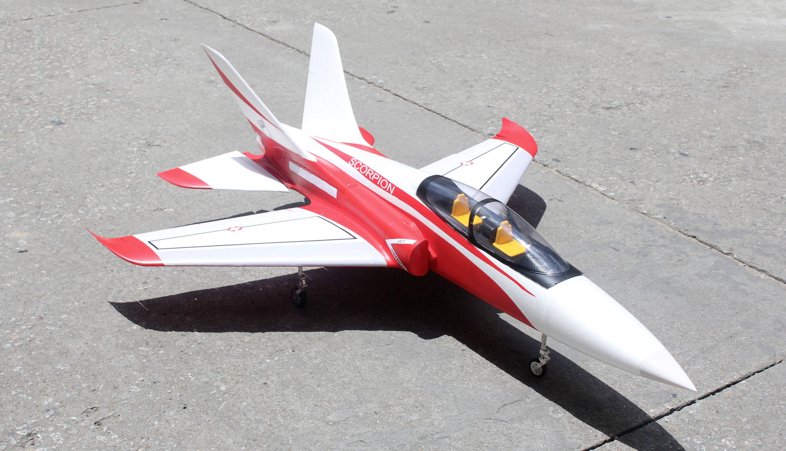 Un modèle d'avion peint en rouge et blanc est posé au sol. Il présente un design élancé et moderne, avec des ailes et des détails de cockpit bien visibles.