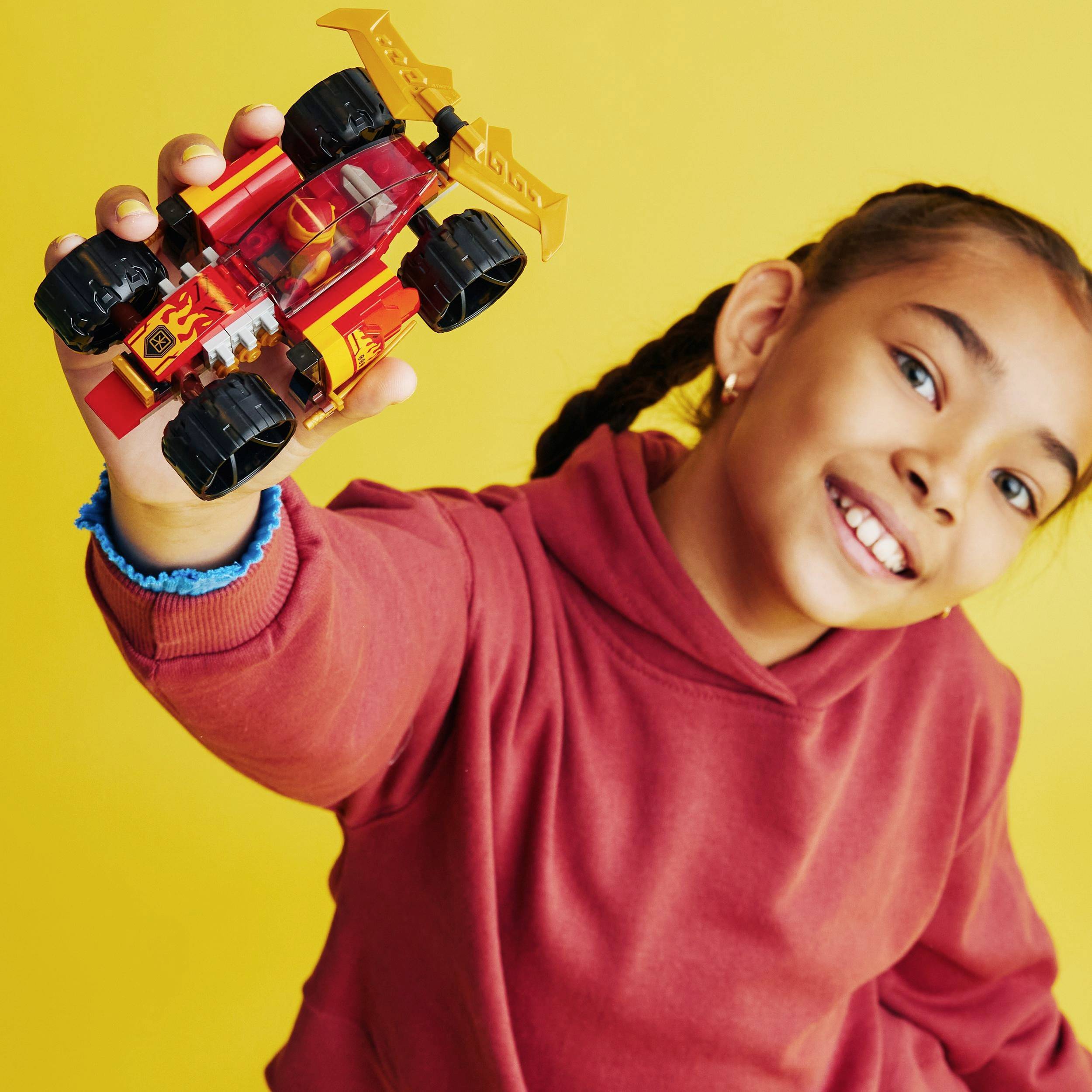 Un enfant tient avec enthousiasme une voiture jouet colorée aux grandes roues, face à l'appareil photo, devant un fond jaune vif.