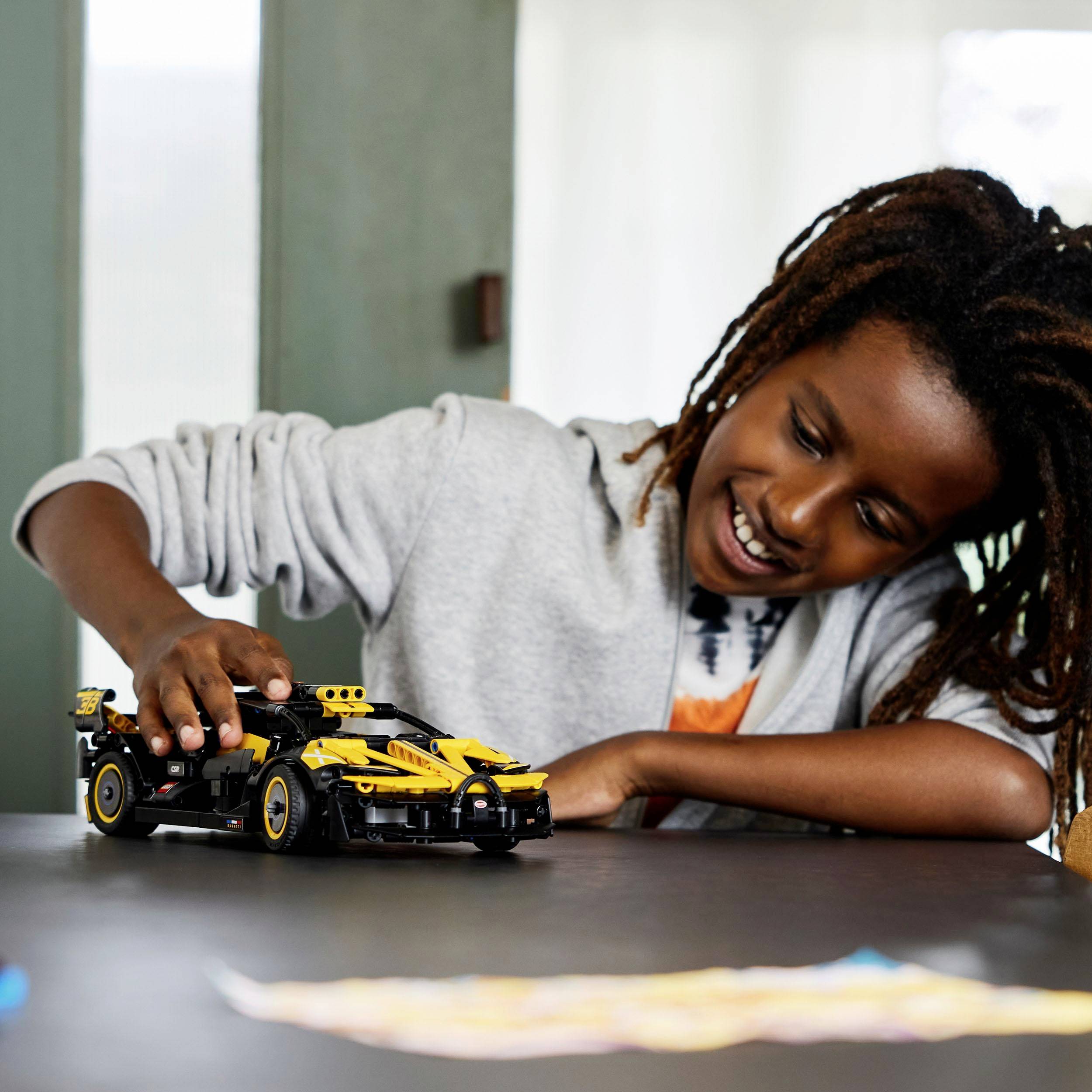 Un enfant joue joyeusement avec une voiture jouet jaune sur une table, dans une pièce avec de grandes fenêtres.