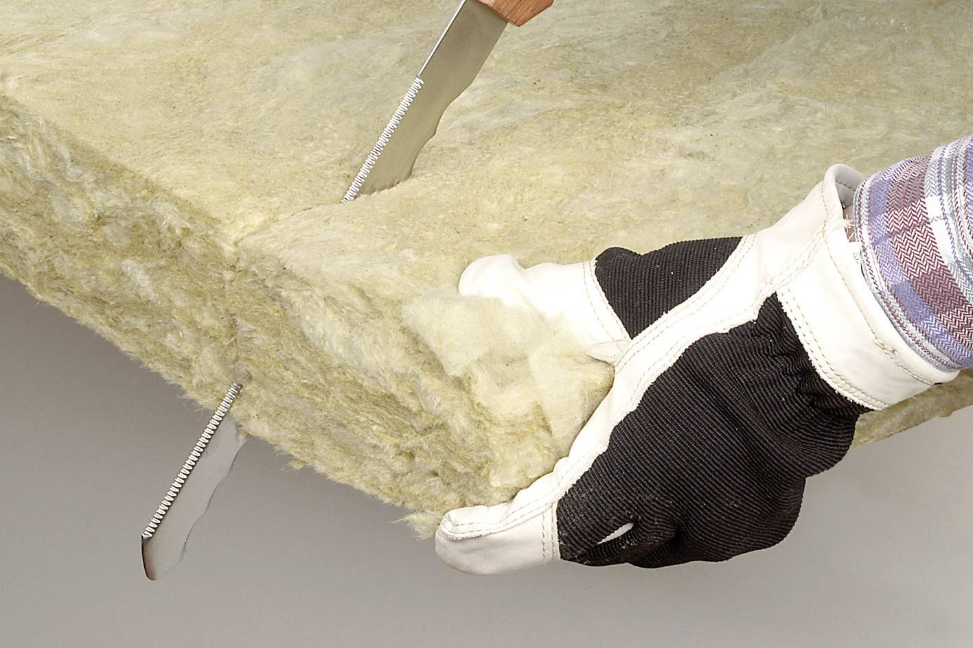 Une personne coupe une plaque isolante en laine minérale à l'aide d'un couteau. Les mains sont protégées par des gants de travail.