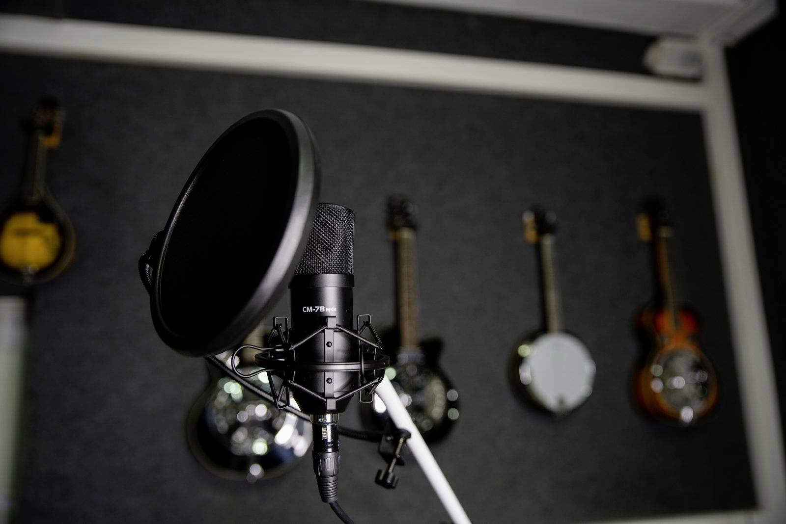 Un microphone de studio avec un filtre anti-pop au premier plan, fixé sur un support. À l'arrière-plan, plusieurs instruments à cordes sont visibles sur le mur.