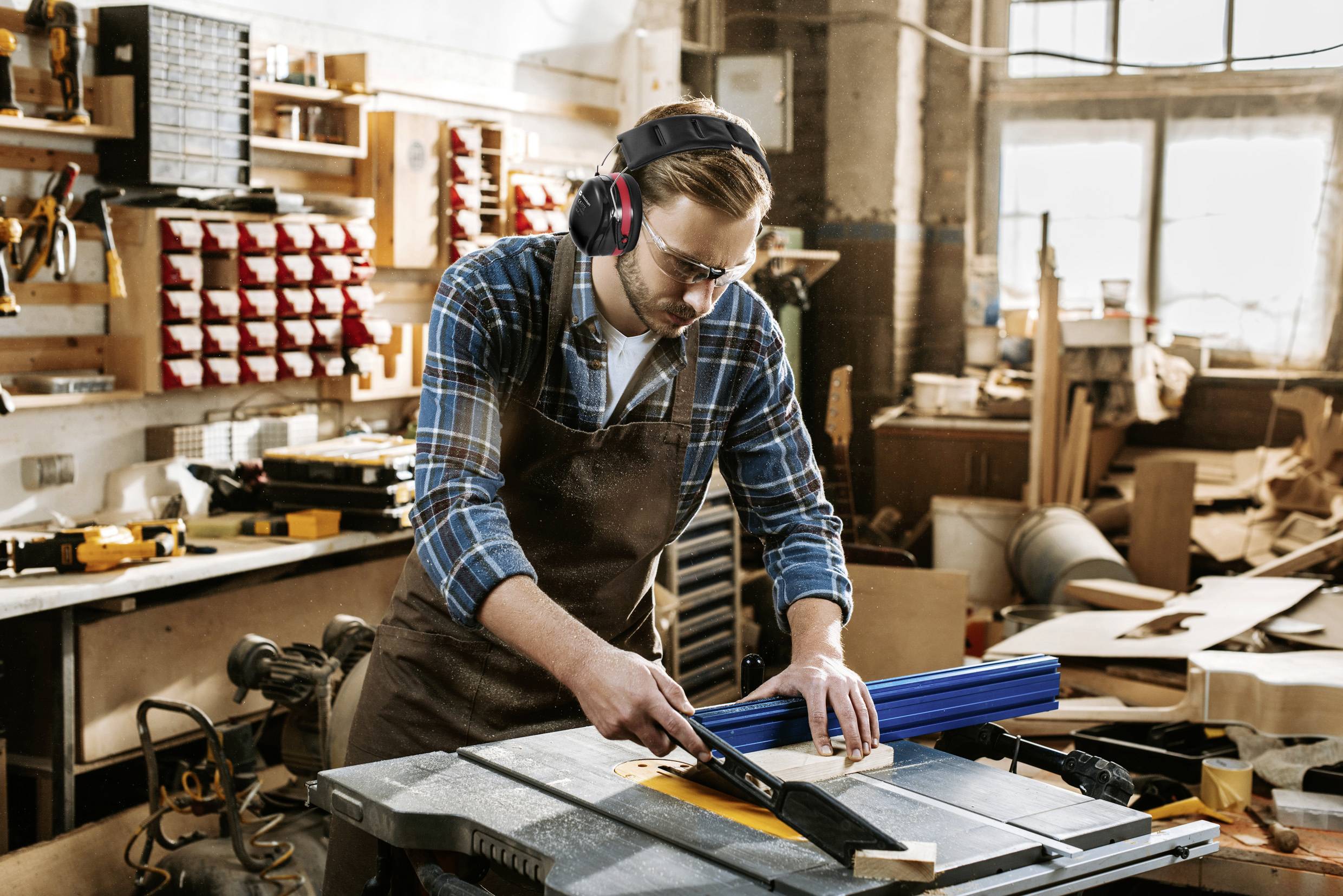Un homme dans un atelier porte des protections auditives et des lunettes de sécurité, tandis qu'il travaille le bois avec une scie circulaire de table.
