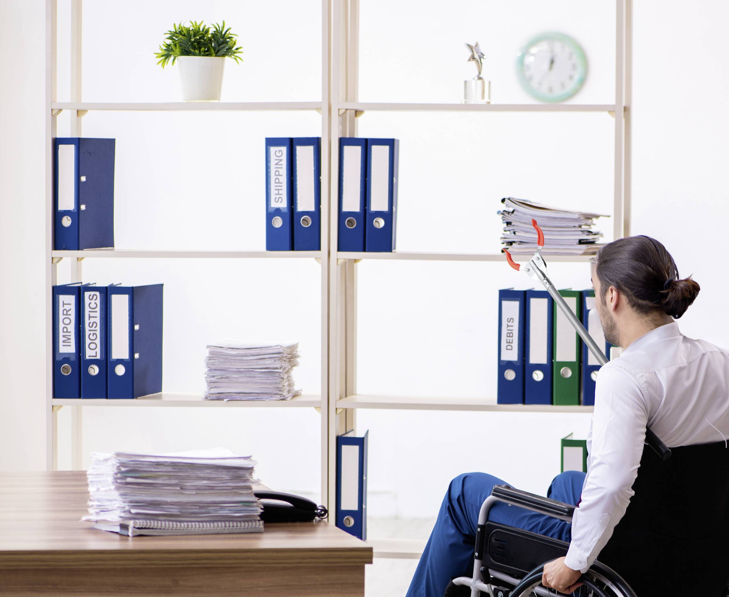 Un homme en fauteuil roulant utilise un bras télescopique pour atteindre un document sur une étagère haute du bureau. Des classeurs sont rangés sur l'étagère.