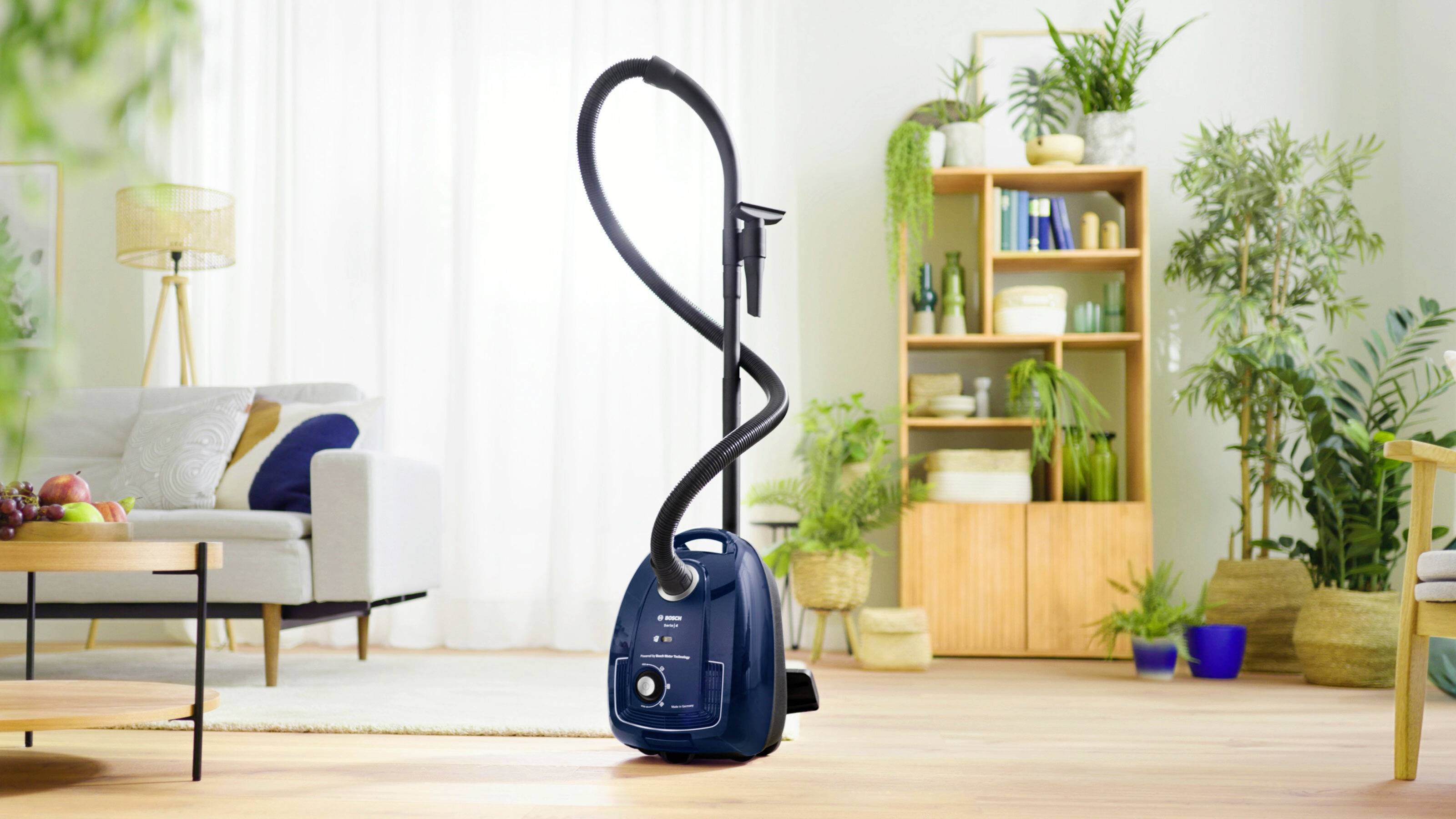 Un aspirateur est placé au centre d'un salon lumineux, avec des plantes, un canapé et une étagère en arrière-plan, prêt à être utilisé.
