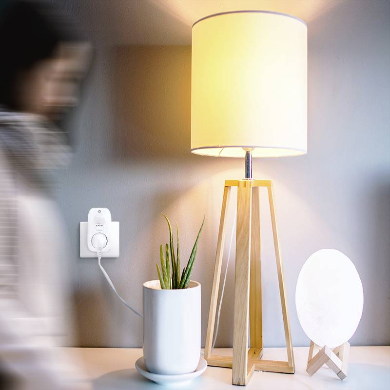 Une table avec une lampe allumée, une plante dans un pot et un objet rond lumineux. Une personne floue passe au premier plan.