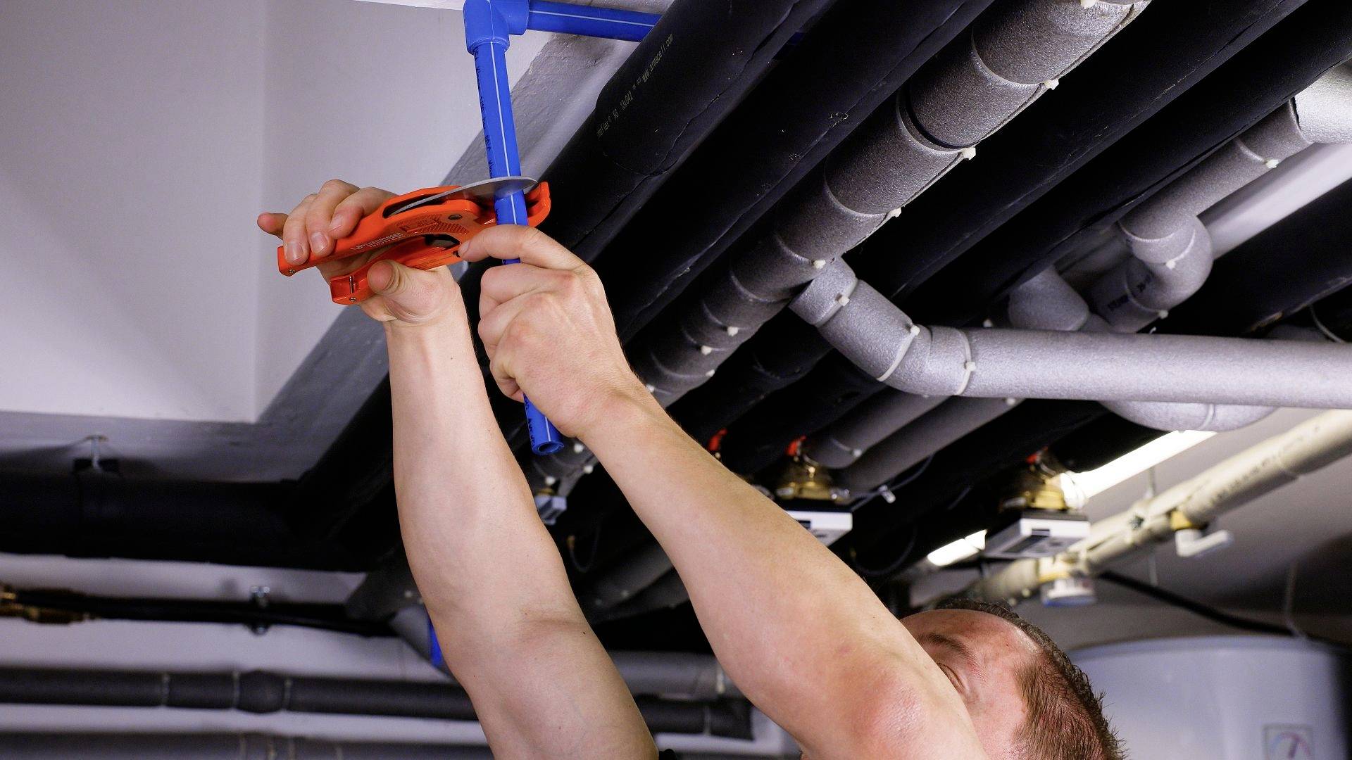 Un artisan répare des canalisations au plafond. Il utilise une pince à tubes orange pour ajuster les raccords.