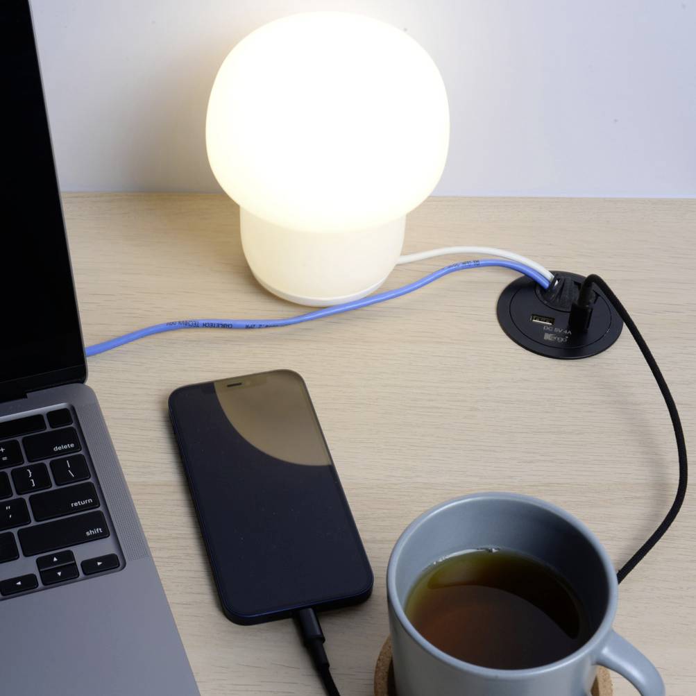 Un bureau avec un ordinateur portable, à côté une lampe, un smartphone et une tasse de thé. Des câbles sont branchés à une prise électrique.