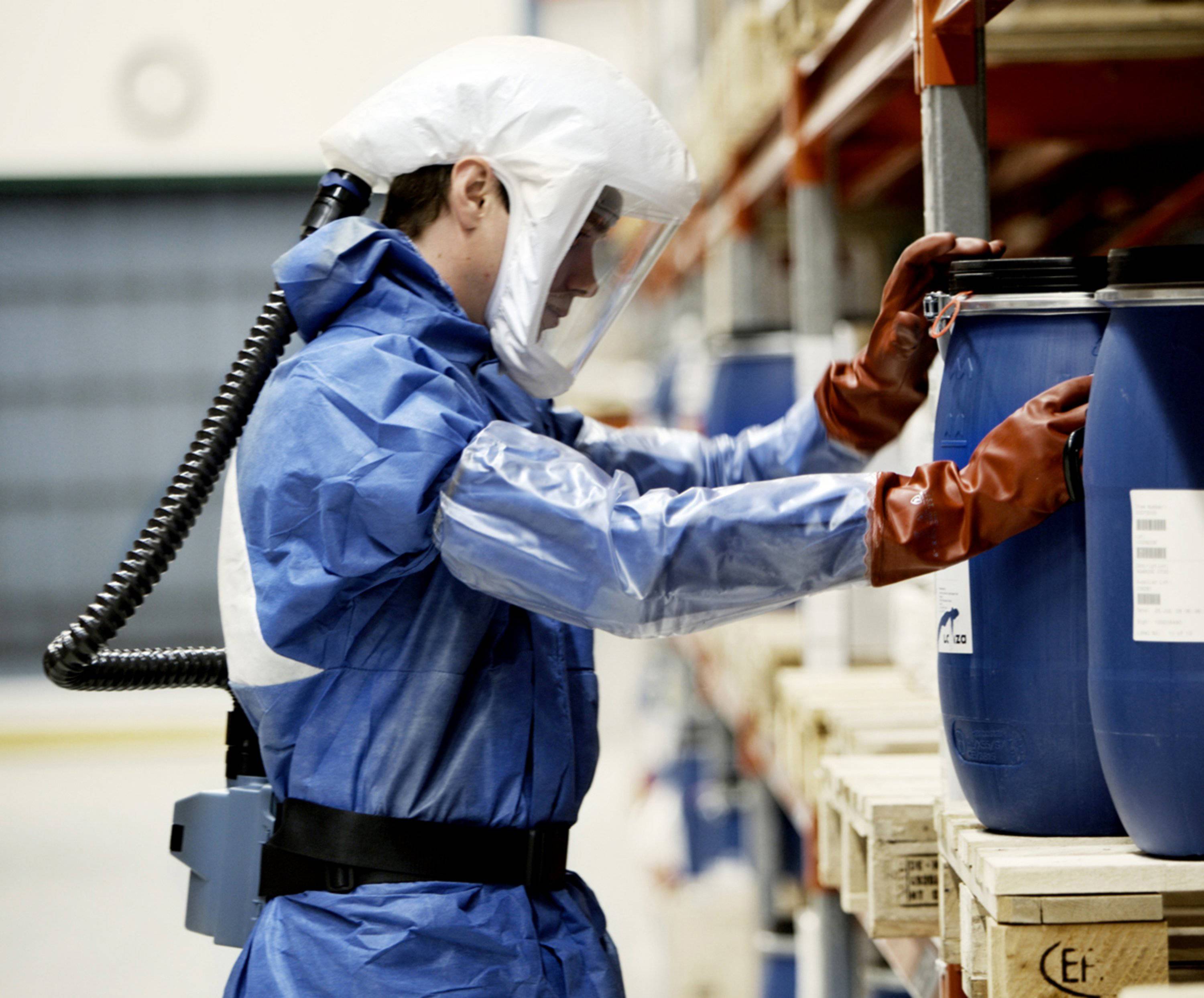 Une personne en combinaison de protection et masque respiratoire inspecte des fûts bleus dans un entrepôt, probablement pour vérifier les normes de sécurité.