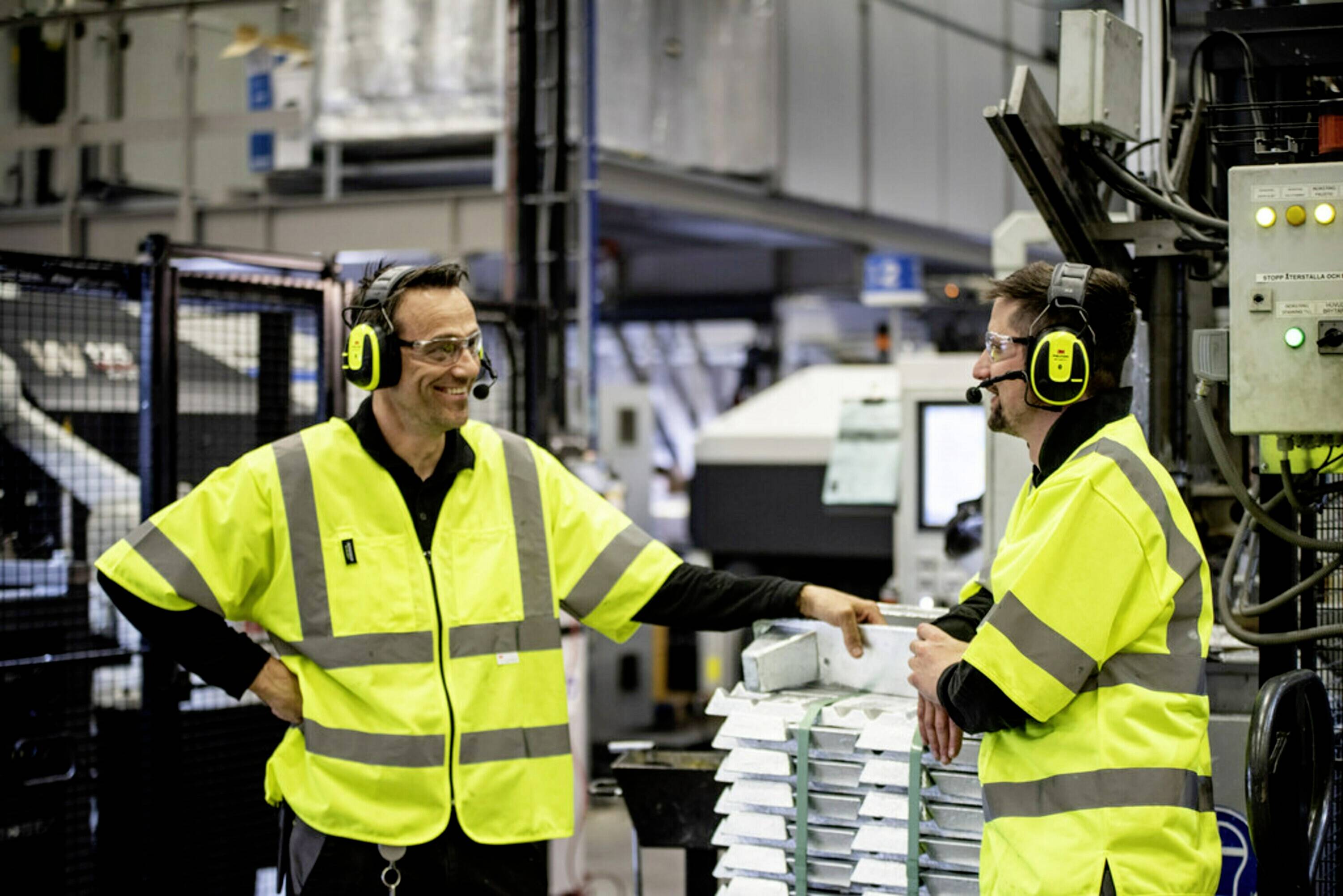 Deux hommes portant des gilets jaunes de signalisation sont debout dans un hall d'usine et discutent. Tous deux portent des protections auditives équipées de microphones.