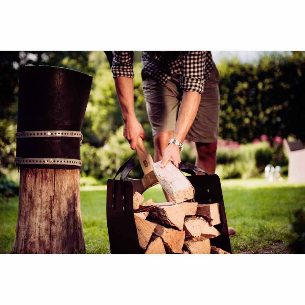 Une personne coupe du bois dans le jardin ; transporte des morceaux de bois vers un panier sombre sur la pelouse. Environnement rural, journée ensoleillée.