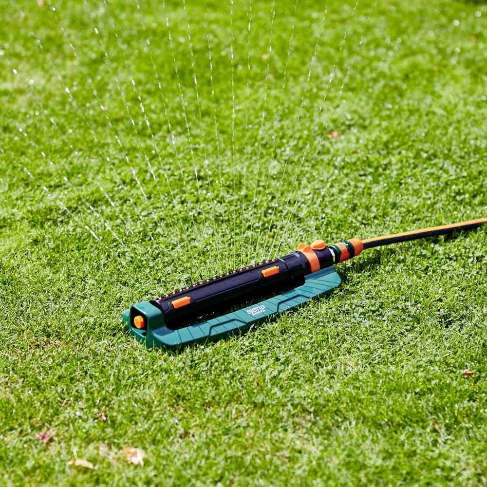 Un arroseur automatique arrose uniformément de l'herbe verte selon un motif régulier. Le tuyau d'arrosage est raccordé et les jets d'eau sont visibles.