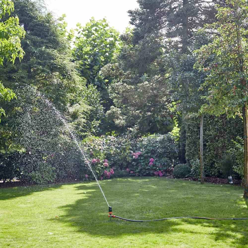 Un arroseur automatique irrigue un jardin ensoleillé et verdoyant, avec des arbres et des arbustes en fleurs en arrière-plan.