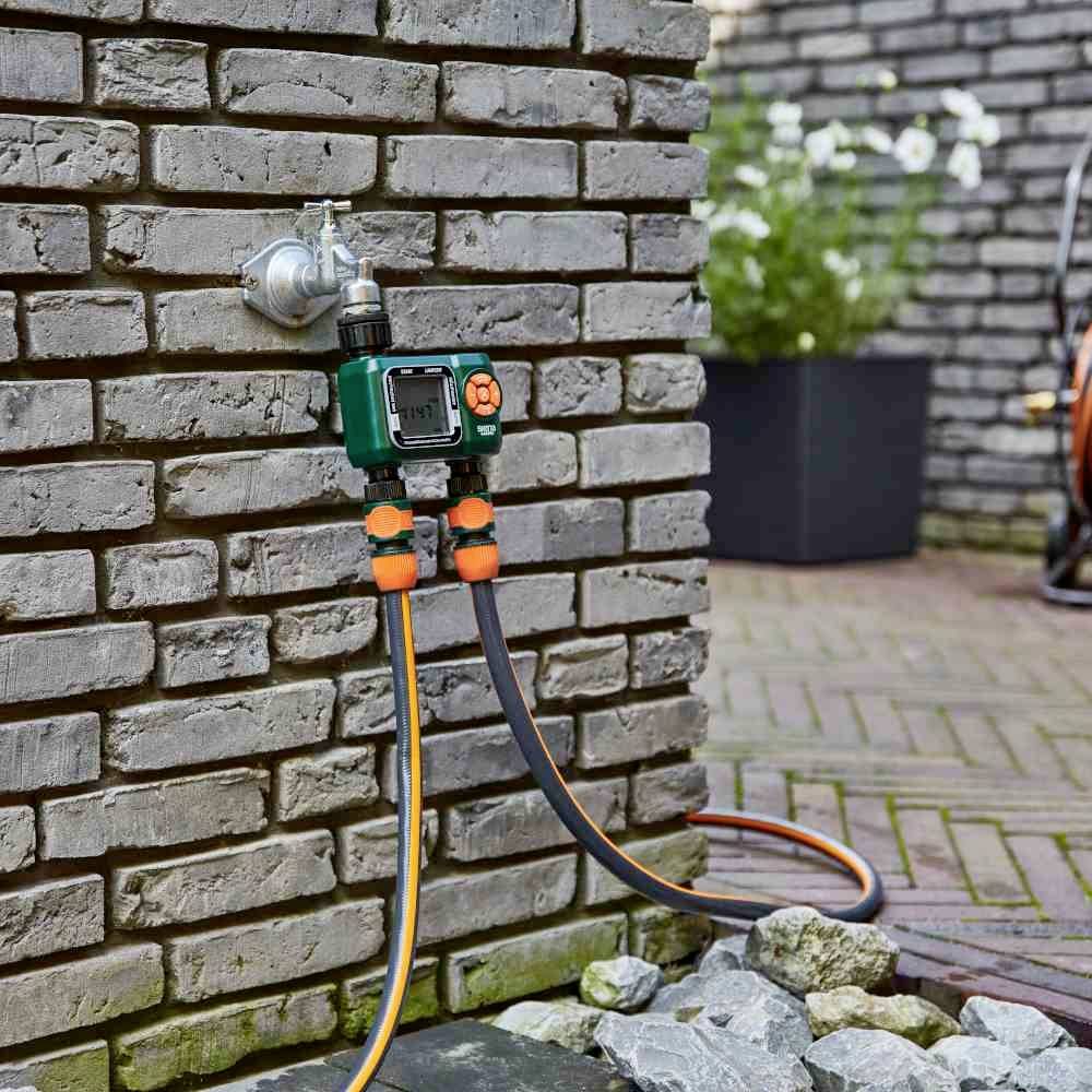 Robinet fixé à un mur de briques avec un système d'irrigation connecté, deux tuyaux. Arrière-plan : terrasse avec des plantes.