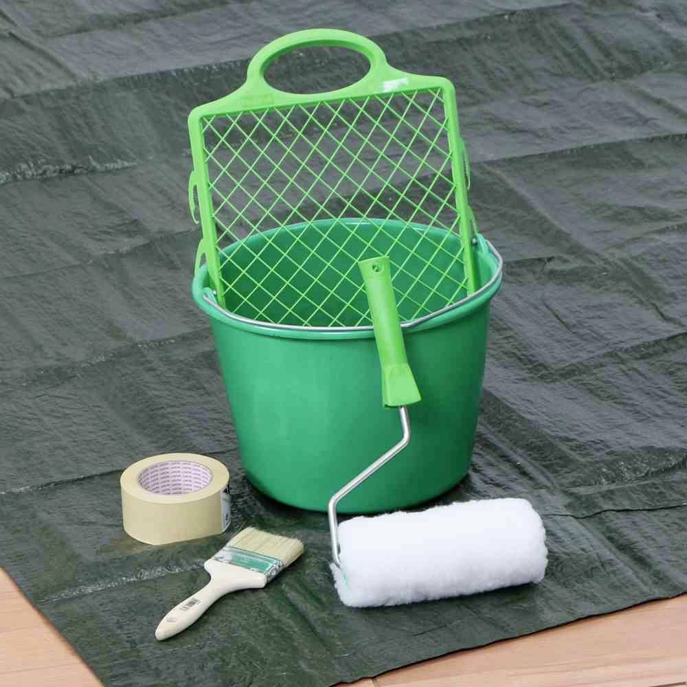 Un seau vert avec un couvercle à tamis, un rouleau à peinture, un pinceau et du ruban adhésif sont posés sur une bâche, prêts pour des travaux de peinture.