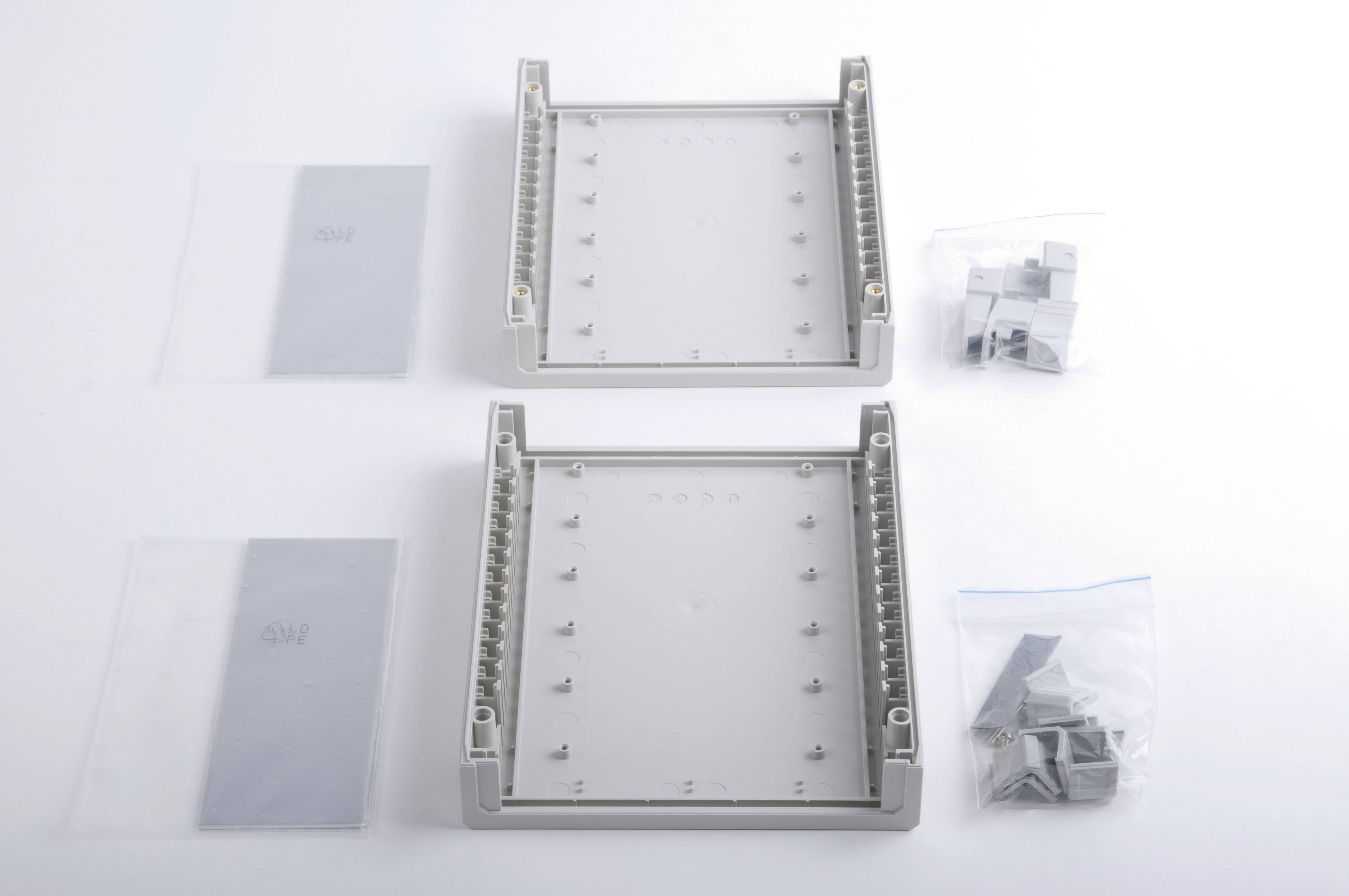 Deux boîtiers en plastique gris sont posés côte à côte, entourés d'emballages contenant des vis et des accessoires de montage sur un fond blanc.
