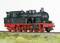 Une locomotive à vapeur noire avec des roues rouges est immobilisée sur des rails. Elle possède une chaudière ronde et de nombreux détails techniques.