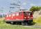 Un train rouge circule sur une voie ferrée étroite à travers un paysage de montagnes verdoyant. Des lignes aériennes et des pylônes électriques bordent la voie.