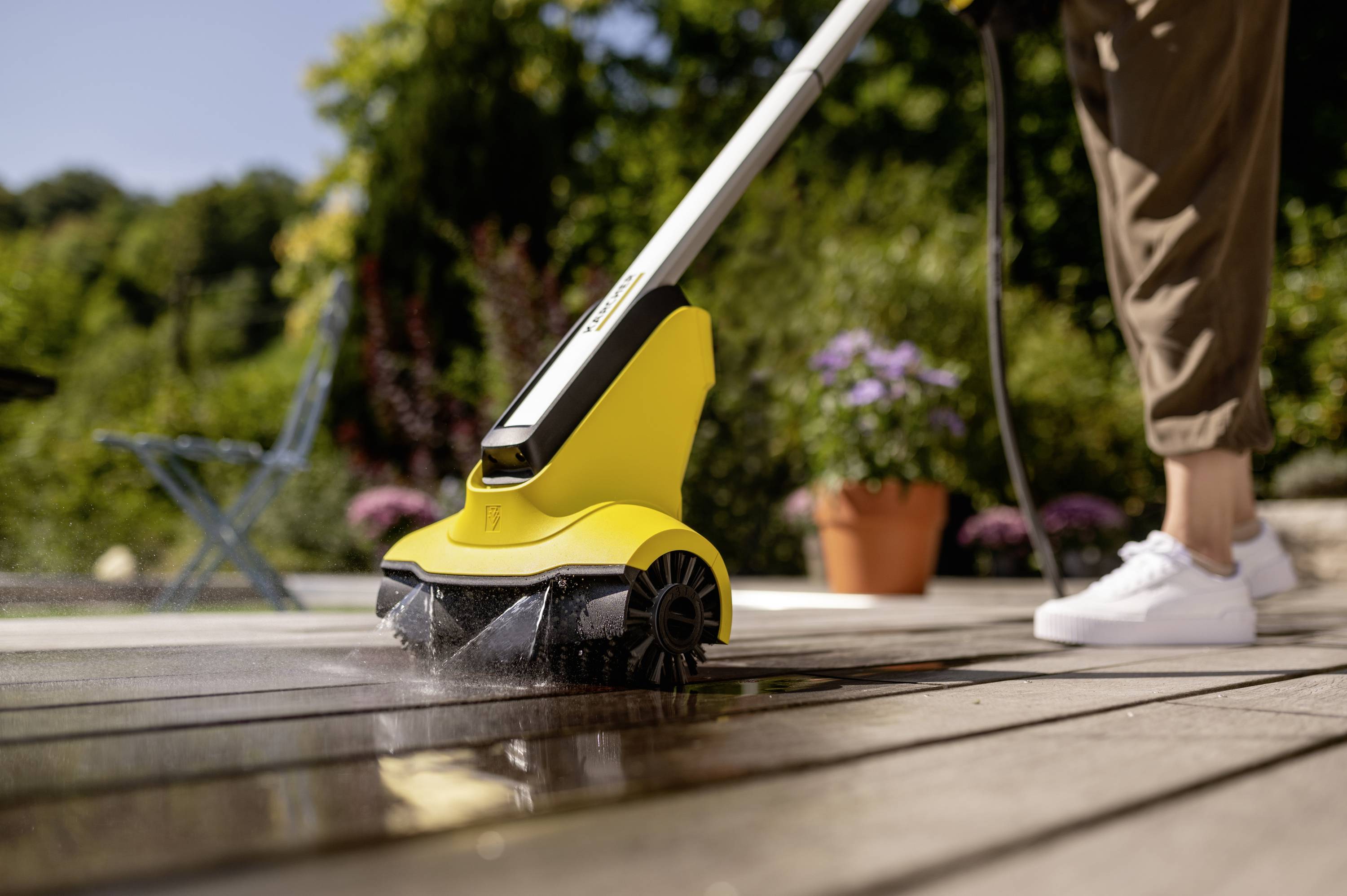 Une personne nettoie une terrasse en bois avec un nettoyeur haute pression jaune par une journée ensoleillée. Des plantes et une chaise bleue sont visibles en arrière-plan.
