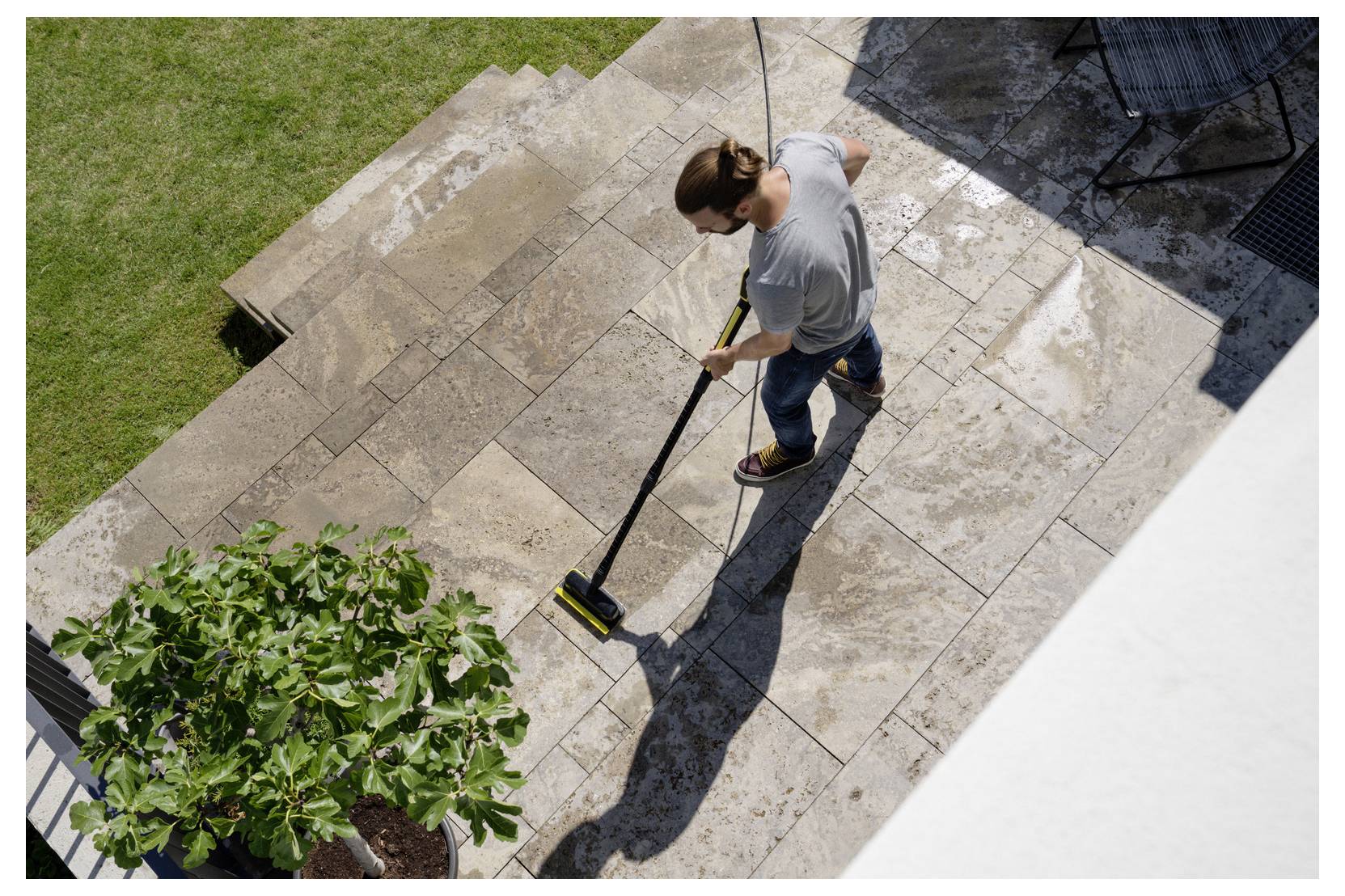 Personne nettoyant une terrasse en pierre avec un balai par une journée ensoleillée, entourée d'herbe et avec un pot de plante visible au premier plan.