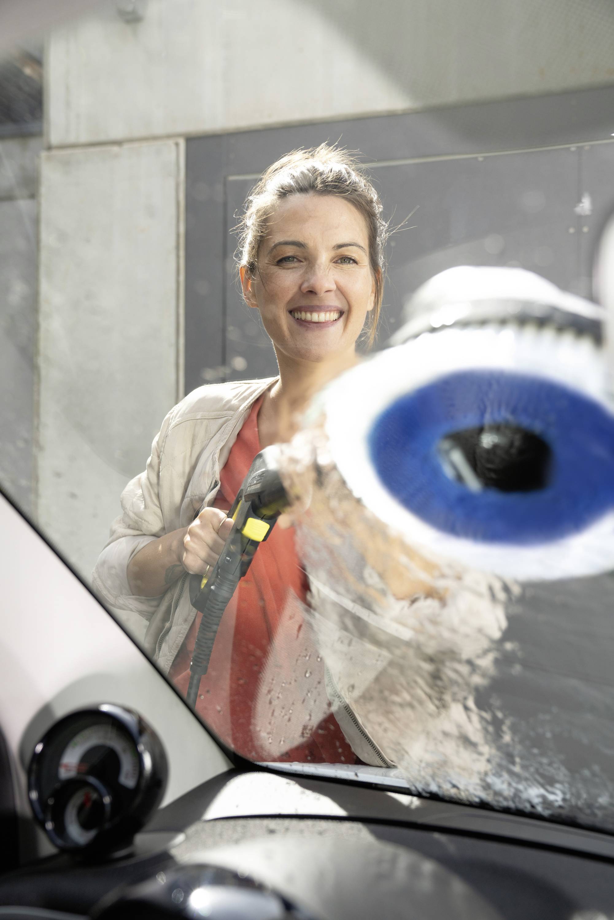 Une personne essuie une vitre de voiture de l'intérieur. Elle sourit et utilise un outil de nettoyage bleu.