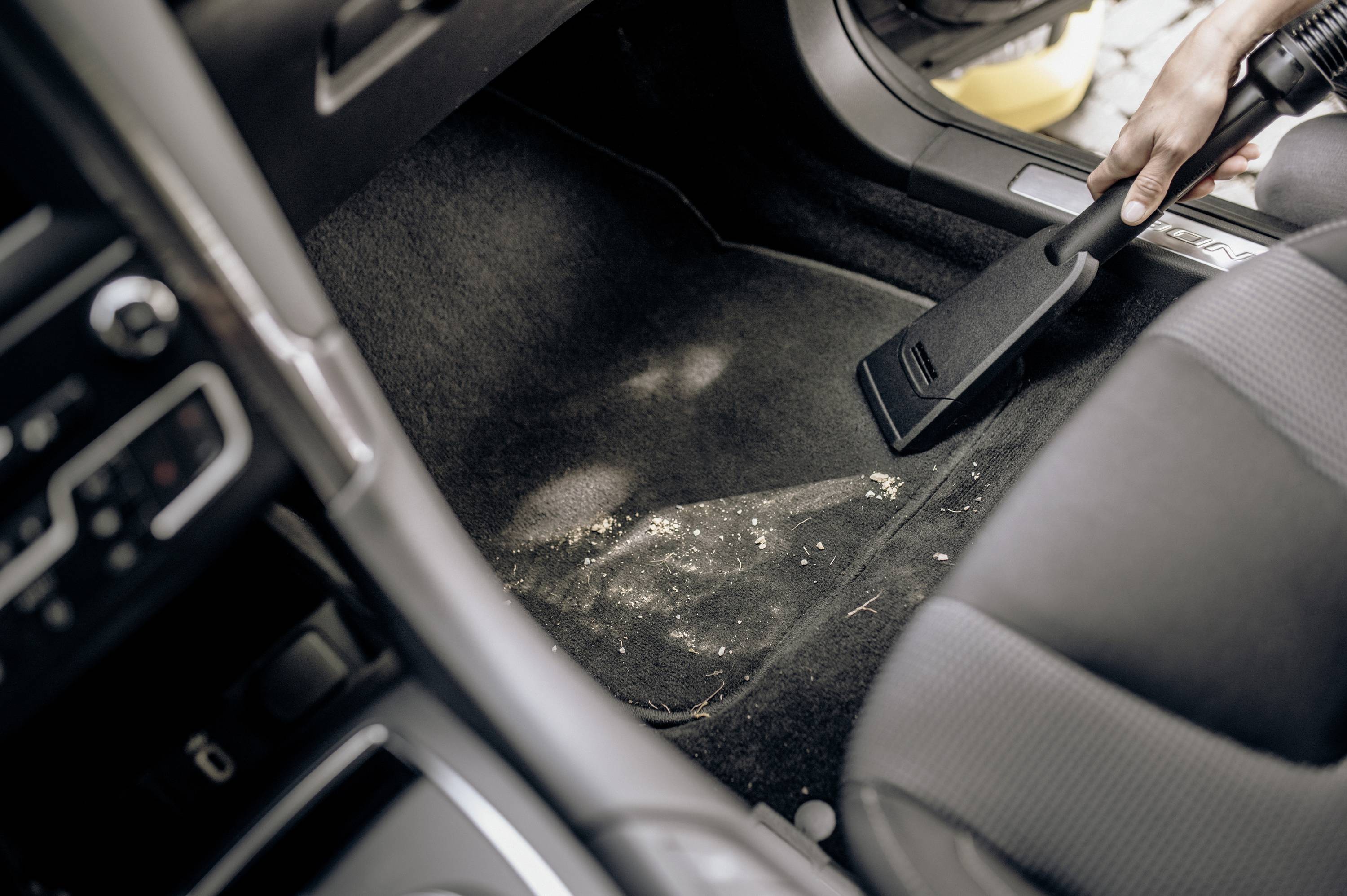 Une personne aspire le sol d'une voiture pour enlever les saletés dispersées. L'aspirateur est équipé d'une buse étroite pour un nettoyage précis.