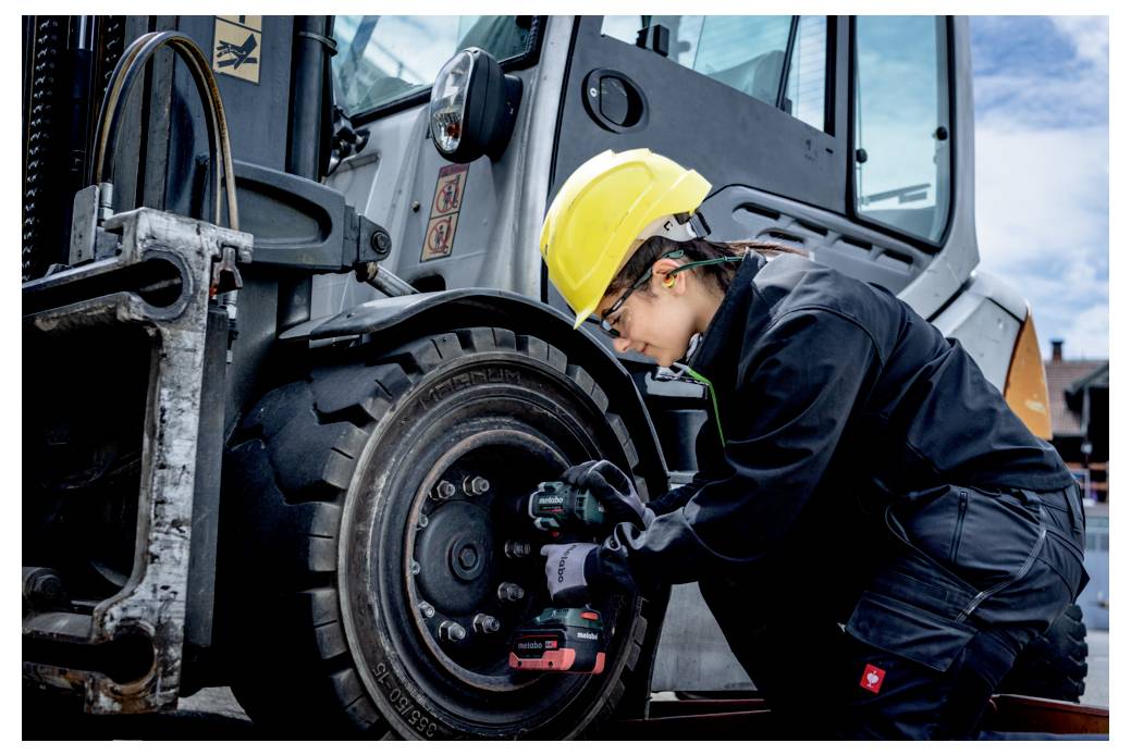 Une personne portant un casque jaune et des vêtements de travail noirs utilise un outil pour serrer les boulons d'un grand pneu de véhicule dans un environnement extérieur.