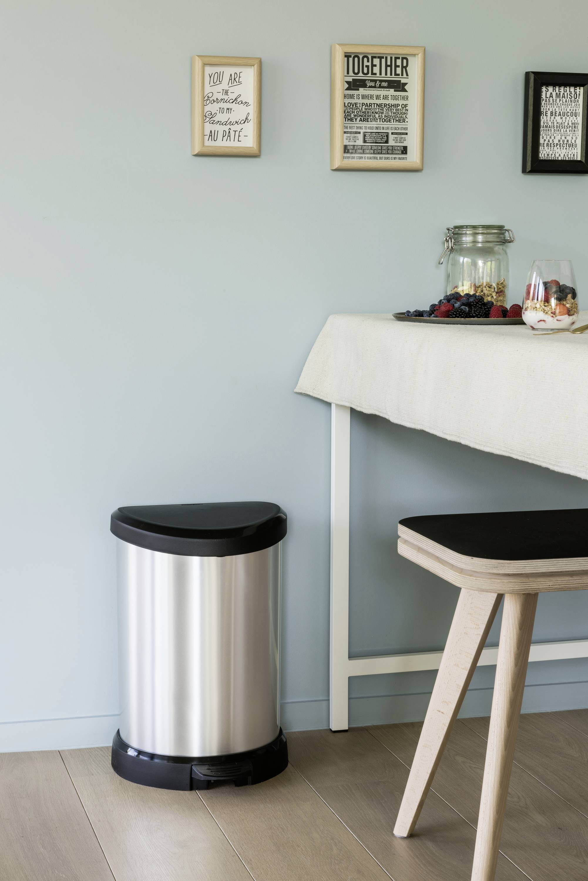 Une poubelle argentée est placée sur un sol en bois à côté d'une table. Sur la table se trouve un bol en verre rempli de fruits, et des images encadrées sont accrochées au mur.