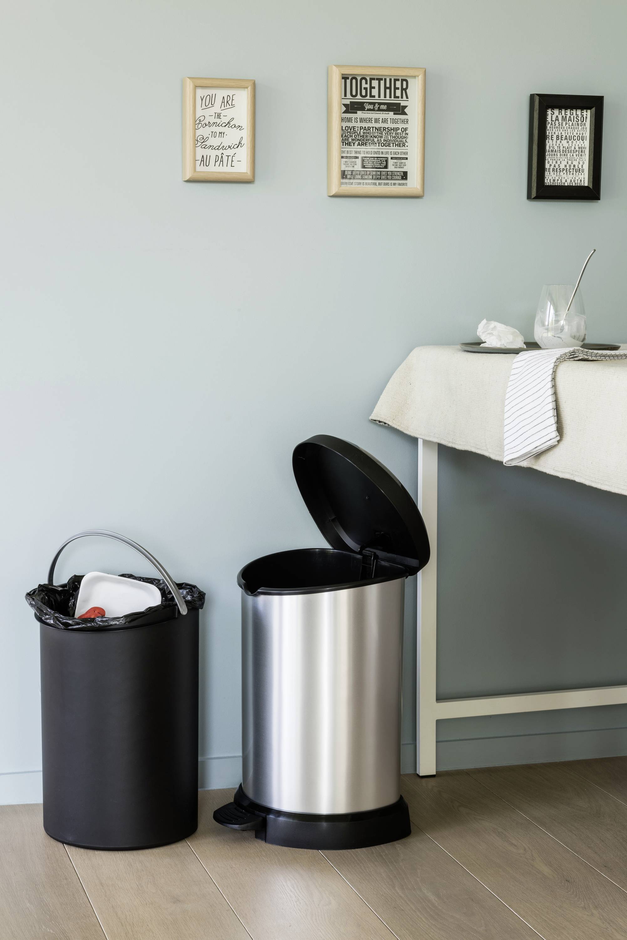 Deux poubelles, l'une noire et l'autre en acier inoxydable, sont côte à côte dans une cuisine, près d'une table avec une assiette et des couverts.