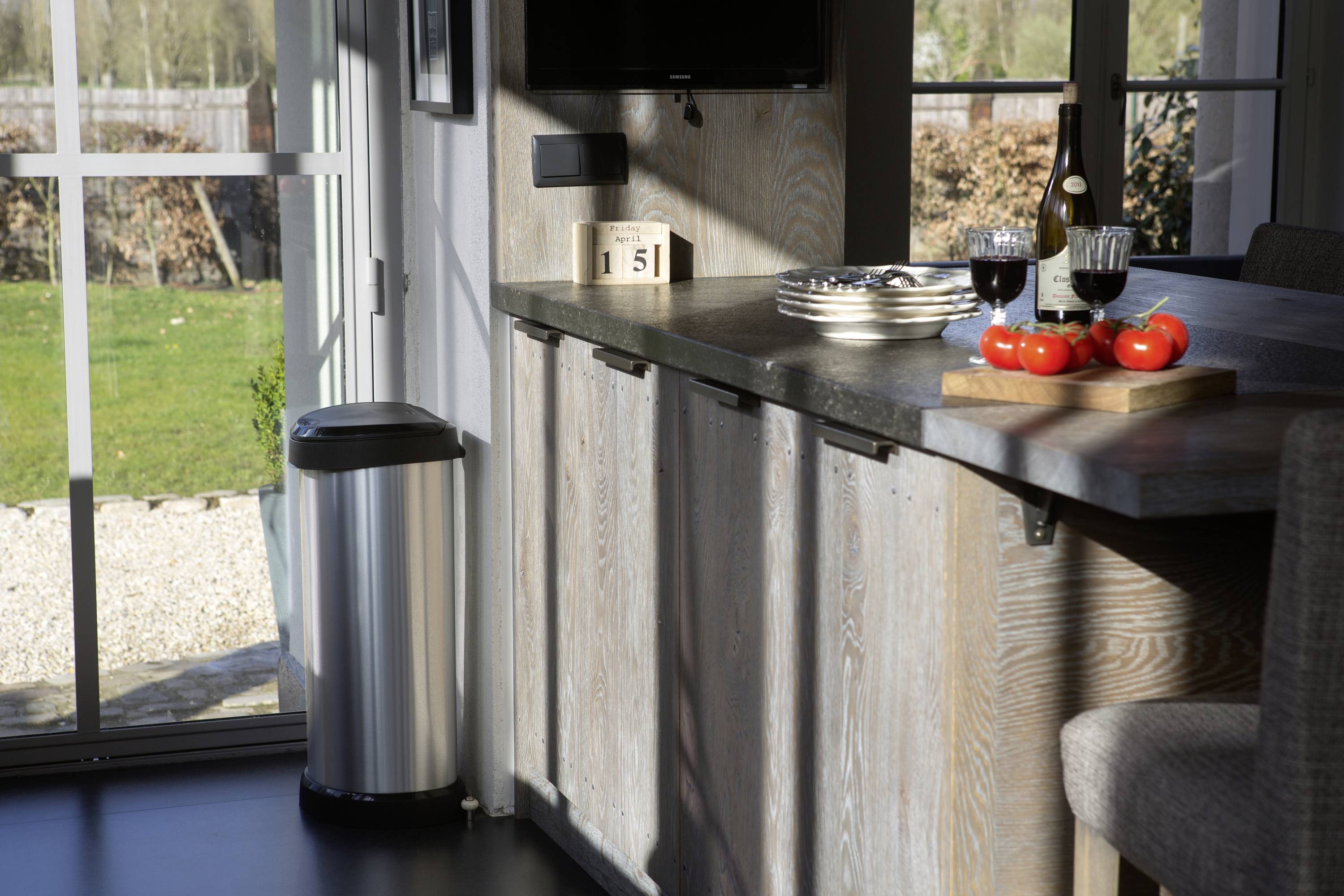 Une cuisine moderne avec un comptoir, de la vaisselle, des tomates et des verres de vin. Dehors, le soleil brille sur un jardin bien entretenu.