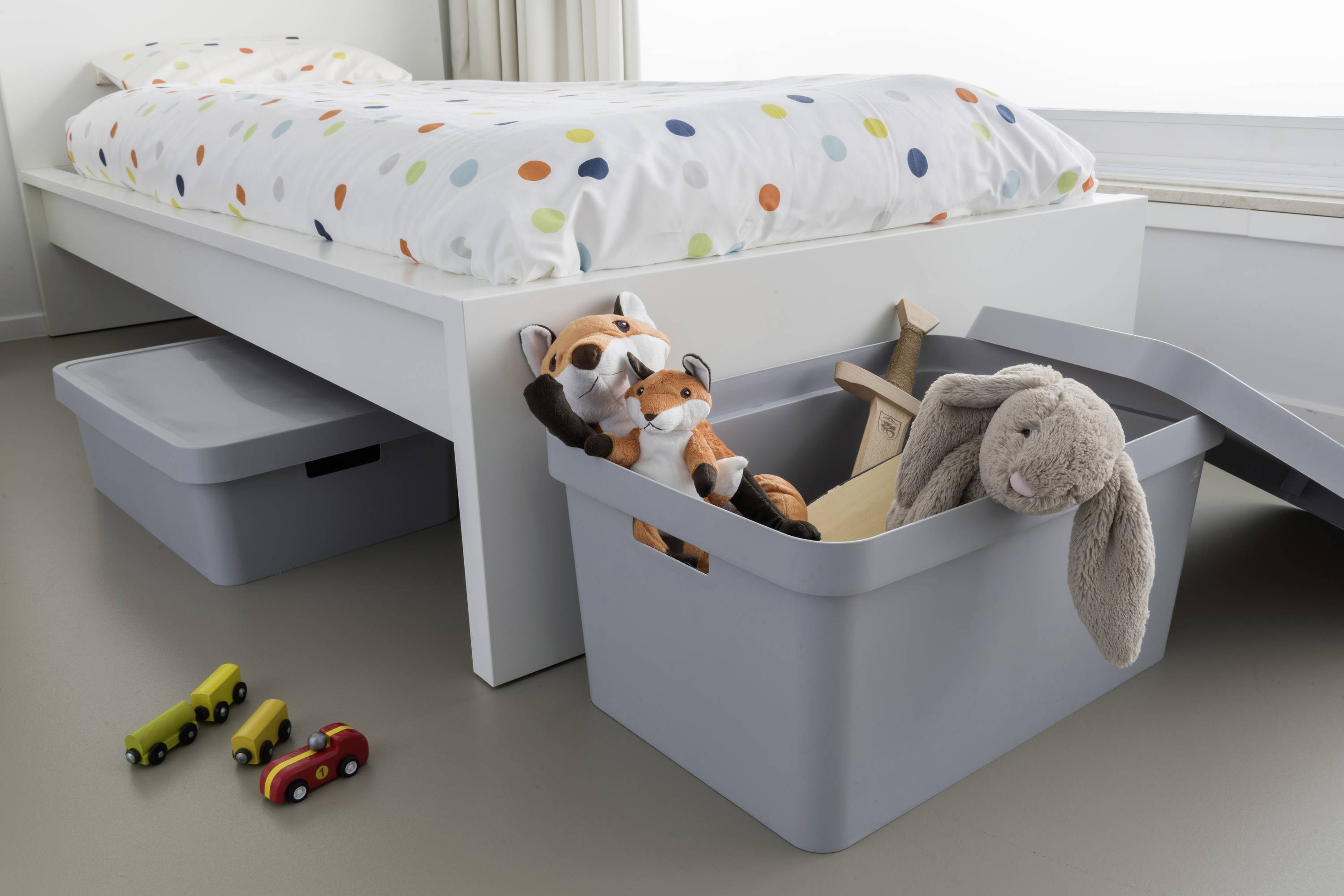 Chambre d'enfant avec un lit blanc. Des voitures miniatures sur le sol. Des boîtes de rangement à côté, remplies de peluches et de jouets en bois.
