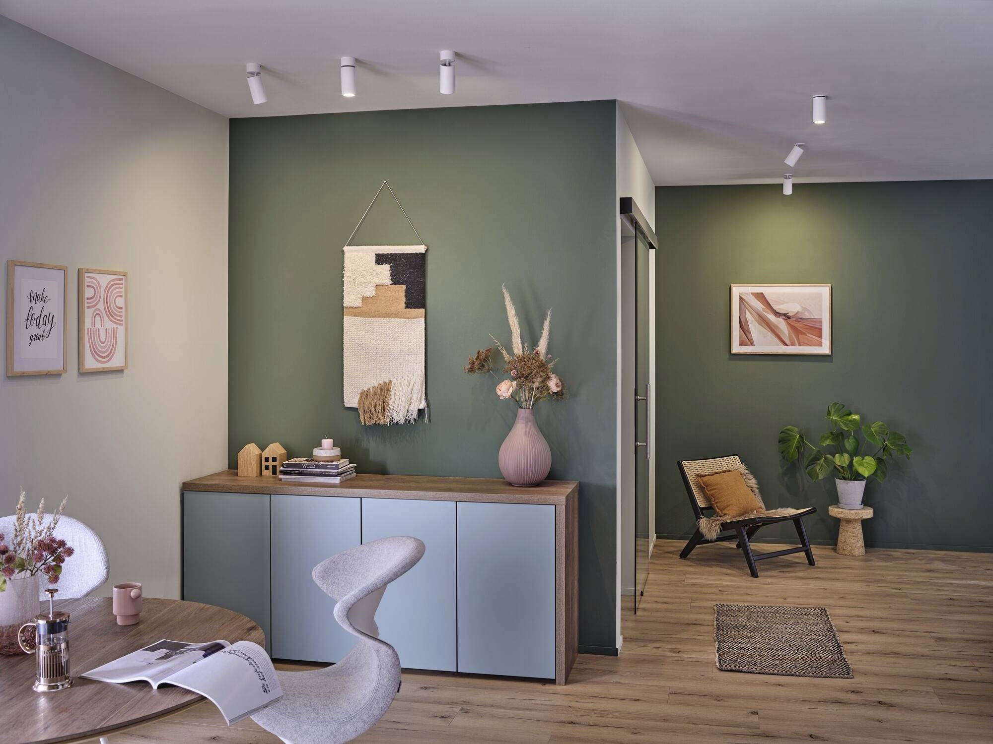 Un salon aménagé de manière moderne avec des murs verts. À gauche, un placard décoré, à droite, une chaise. Des sols en bois chaleureux.