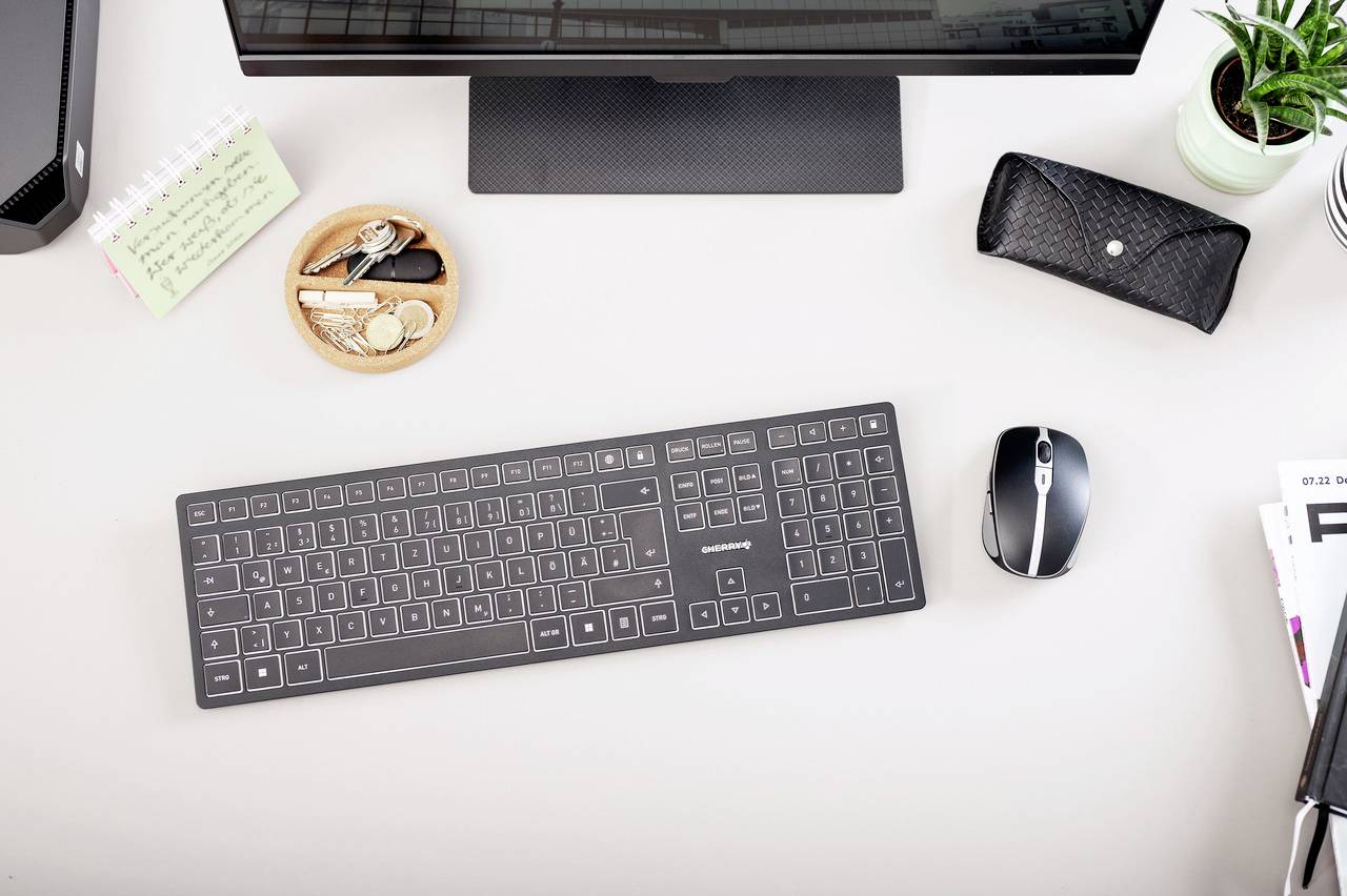 Bureau avec ordinateur, moniteur, clavier, souris, carnet de notes, clés, lunettes dans leur étui et plante. Espace de travail organisé.