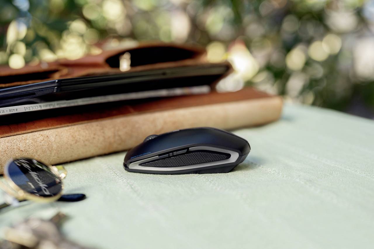 Souris d'ordinateur sans fil sur une table verte, devant un cahier brun et un ordinateur portable. L'éclairage suggère une prise de vue en extérieur.