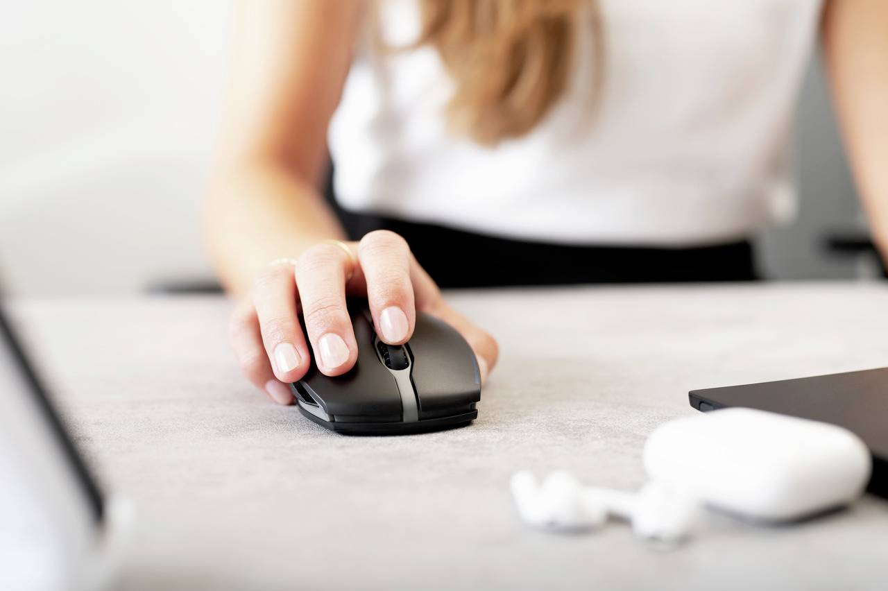 Une personne portant un haut blanc utilise une souris d'ordinateur noire sur un bureau. Des écouteurs sans fil sont visibles au premier plan.