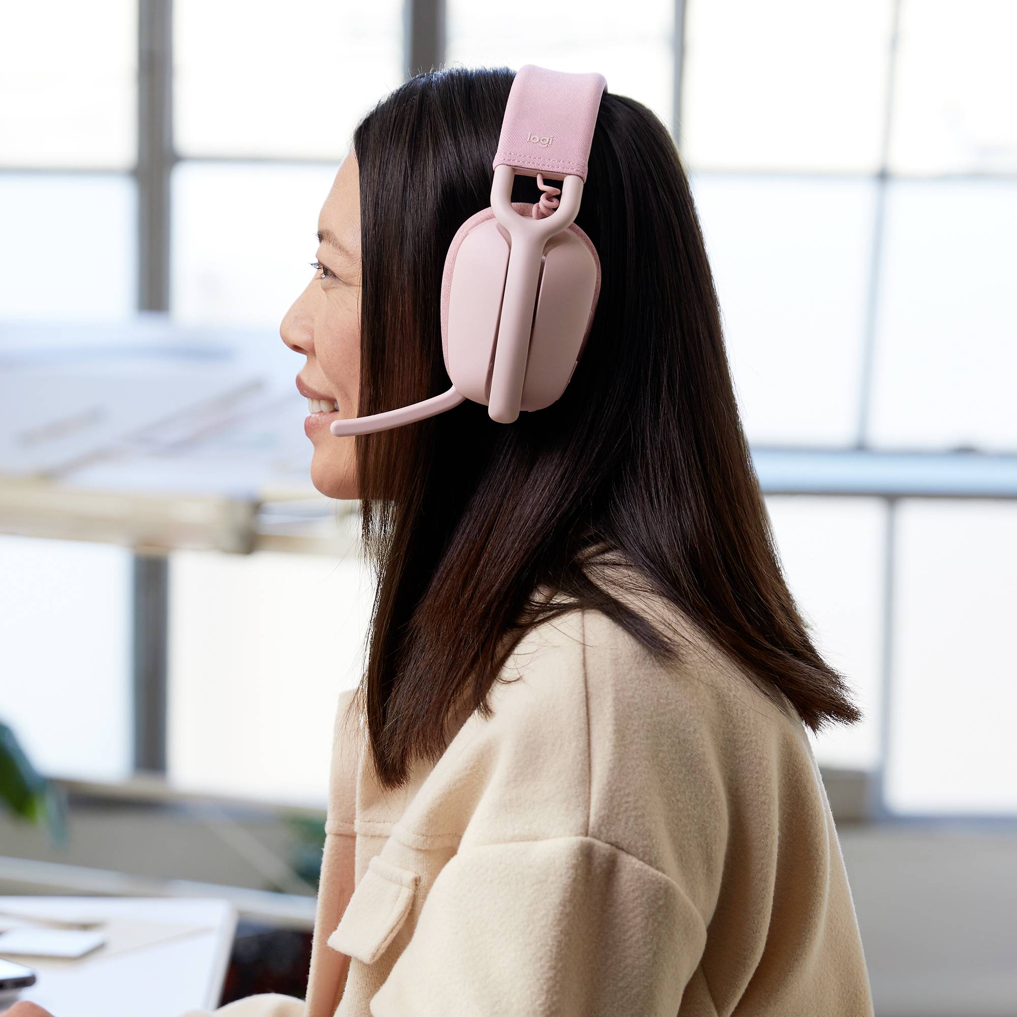 Une femme aux cheveux longs porte des écouteurs roses avec microphone et est assise à un bureau. Une fenêtre est visible en arrière-plan.