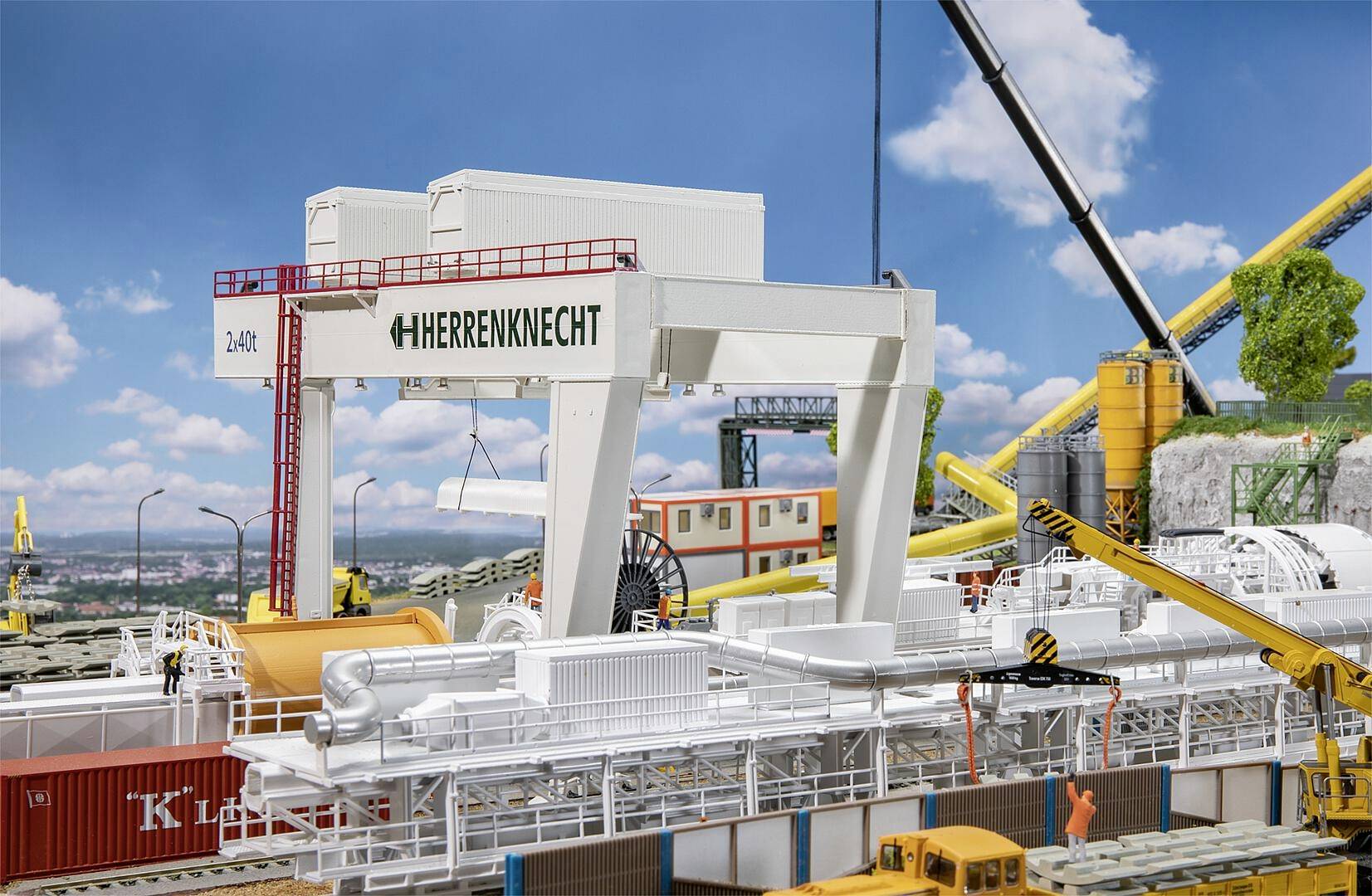 Installation de convoyeur avec une grue et des équipements de construction sur un terrain industriel, entourée de conteneurs et de machines ; ciel bleu en arrière-plan.