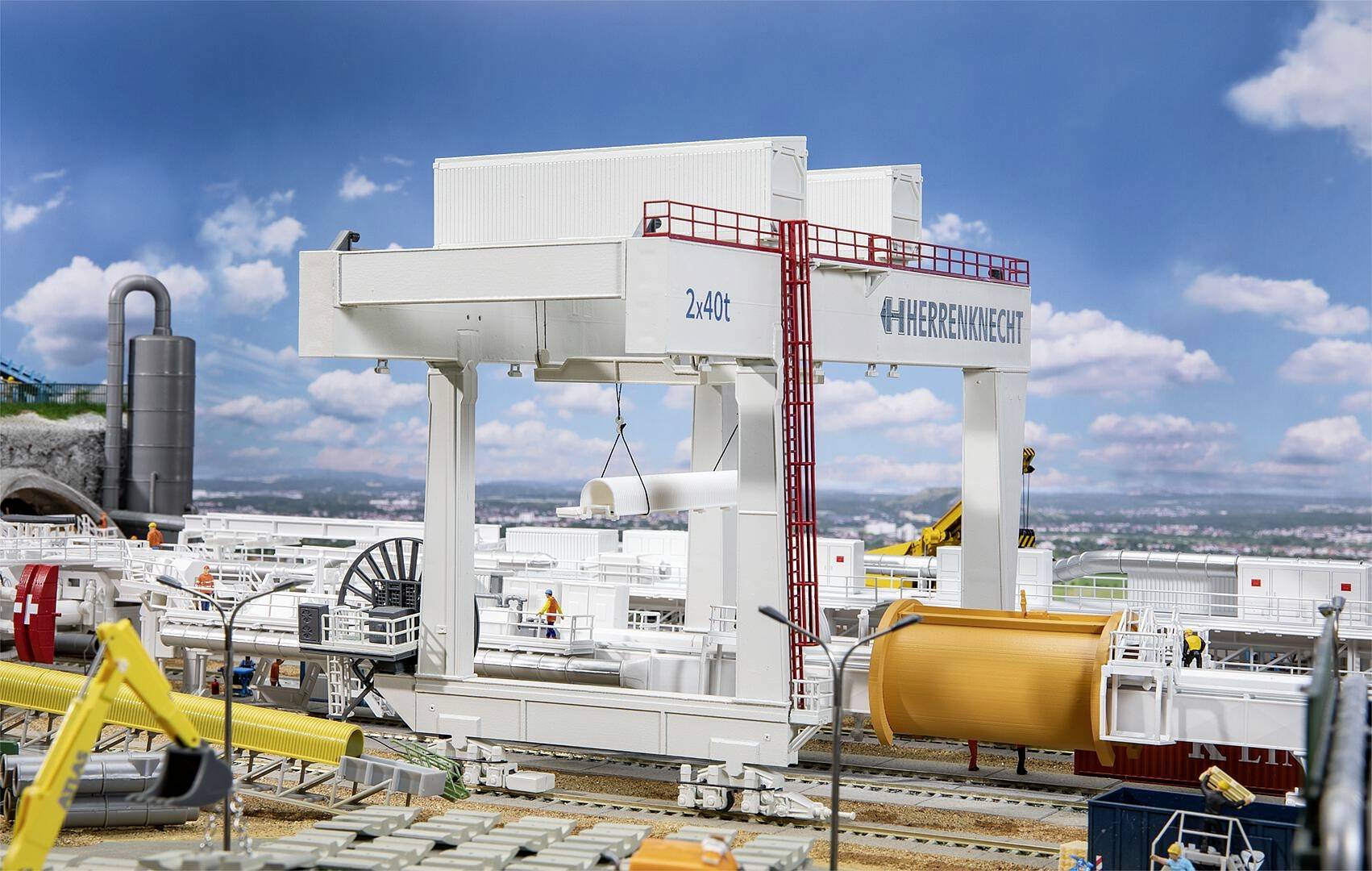 Une grande grue soulève un élément de construction dans une installation industrielle. À l'arrière-plan, on peut voir différentes machines et équipements.