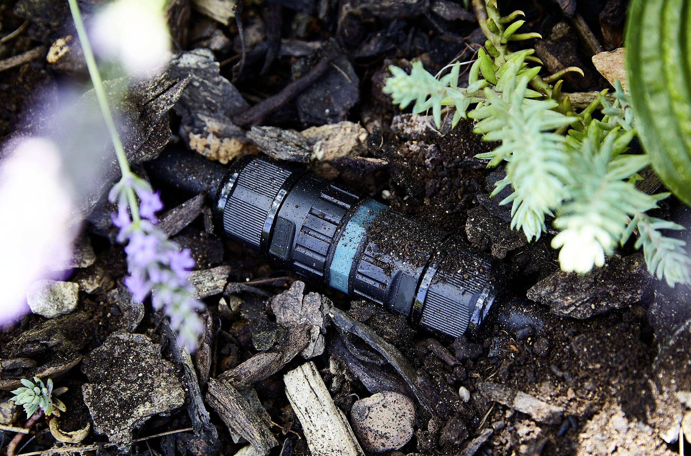 Un connecteur de tuyau d'arrosage noir enfoui dans la terre entre des plantes et du paillis d'écorce.