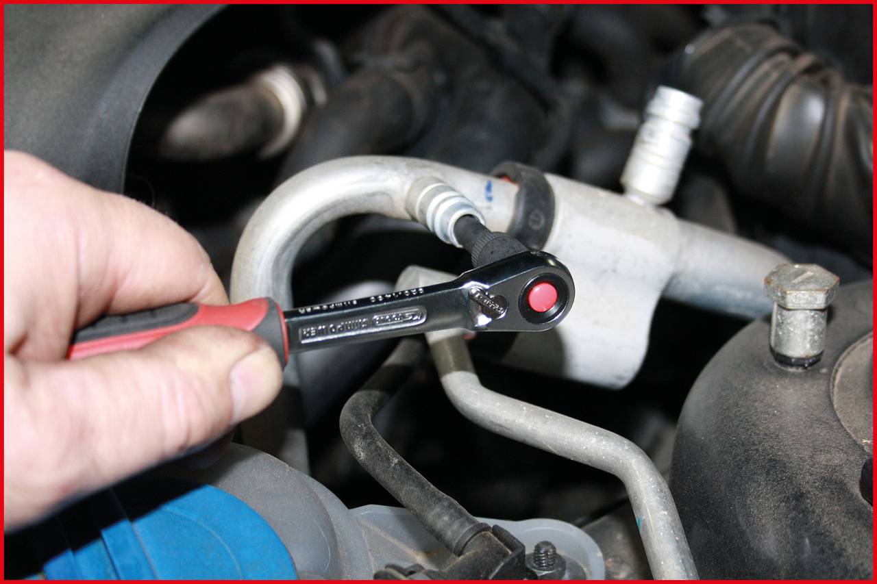 Une main tient une clé à cliquet et serre une vis sur une pièce automobile, probablement dans le compartiment moteur.