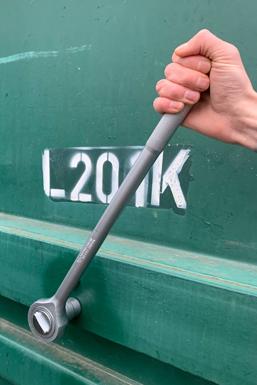 Une main tient une clé à molette devant un fond vert. L'arrière-plan présente le marquage peint 'L-201K'.