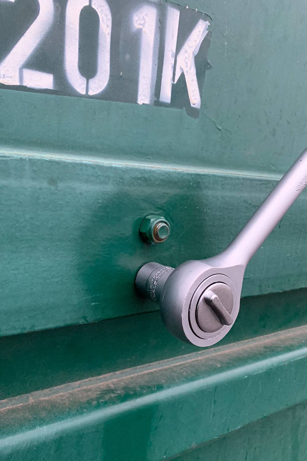 Une clé à molette serre un écrou sur une surface métallique verte. Une partie du chiffre '201K' est visible en haut.