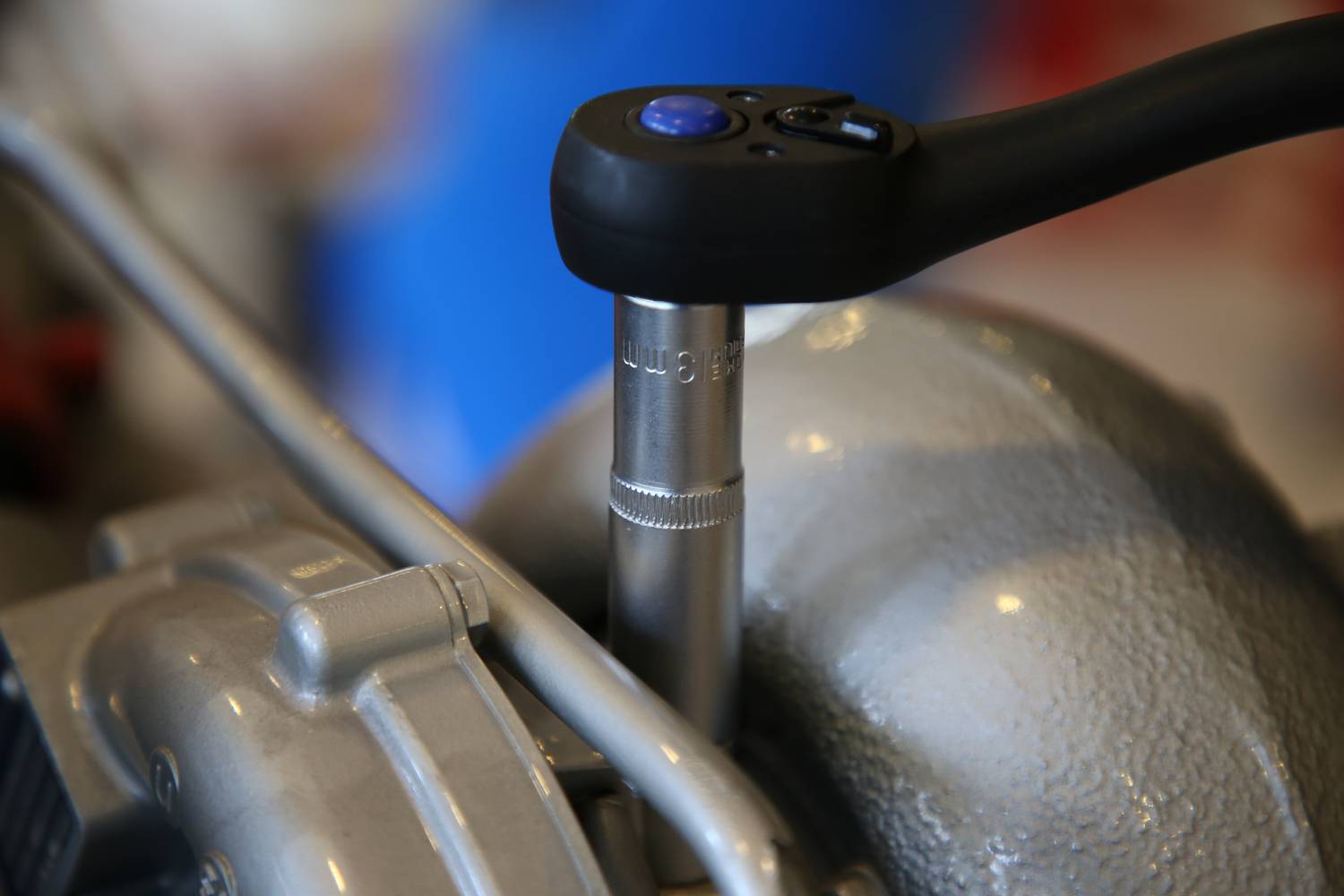 Une clé dynamométrique serre une vis sur une pièce métallique de moteur. En arrière-plan, des surfaces floues bleues et rouges.